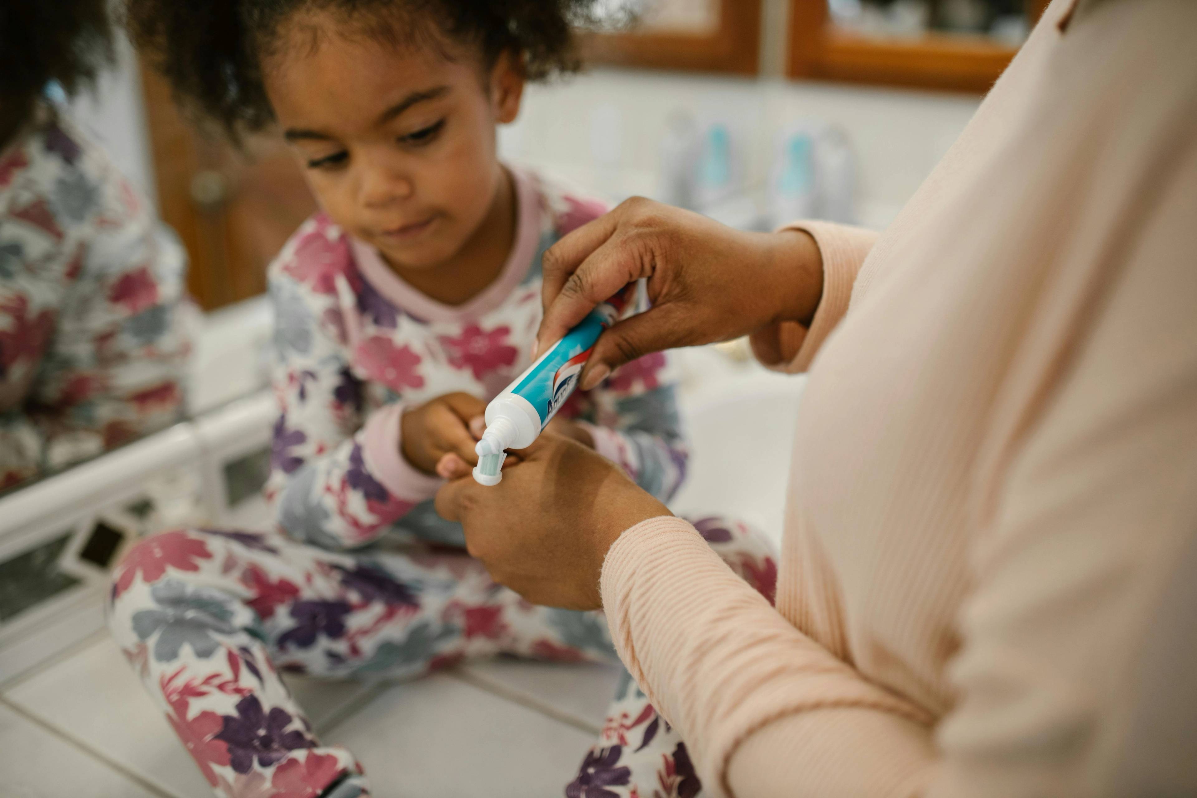 child sat wearing pyjamas holding toothbrush while parent squeezes toothpaste tube