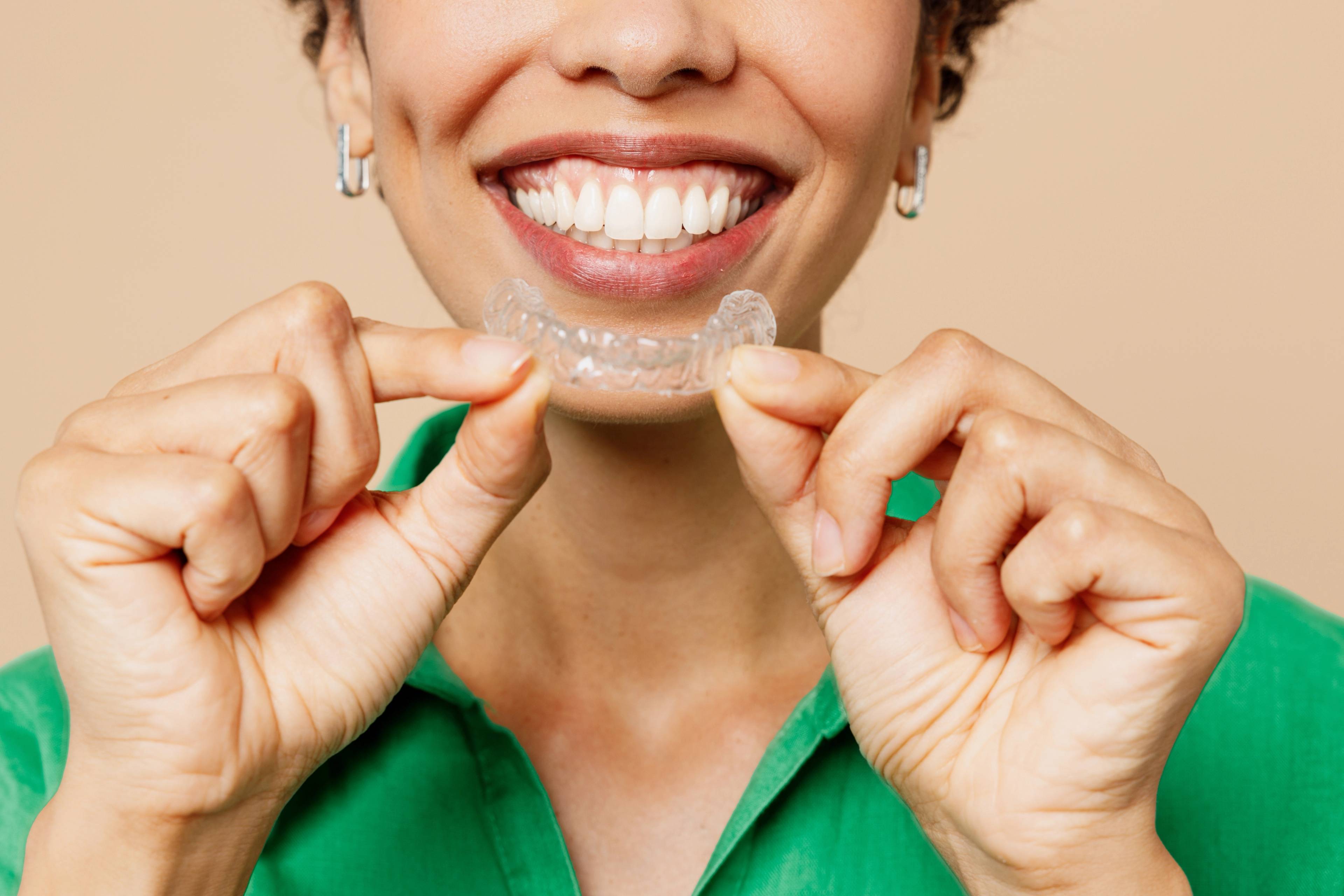 lower half of women's face smiling and holding a clear aligner tray, wearing a green shirt on a beige background