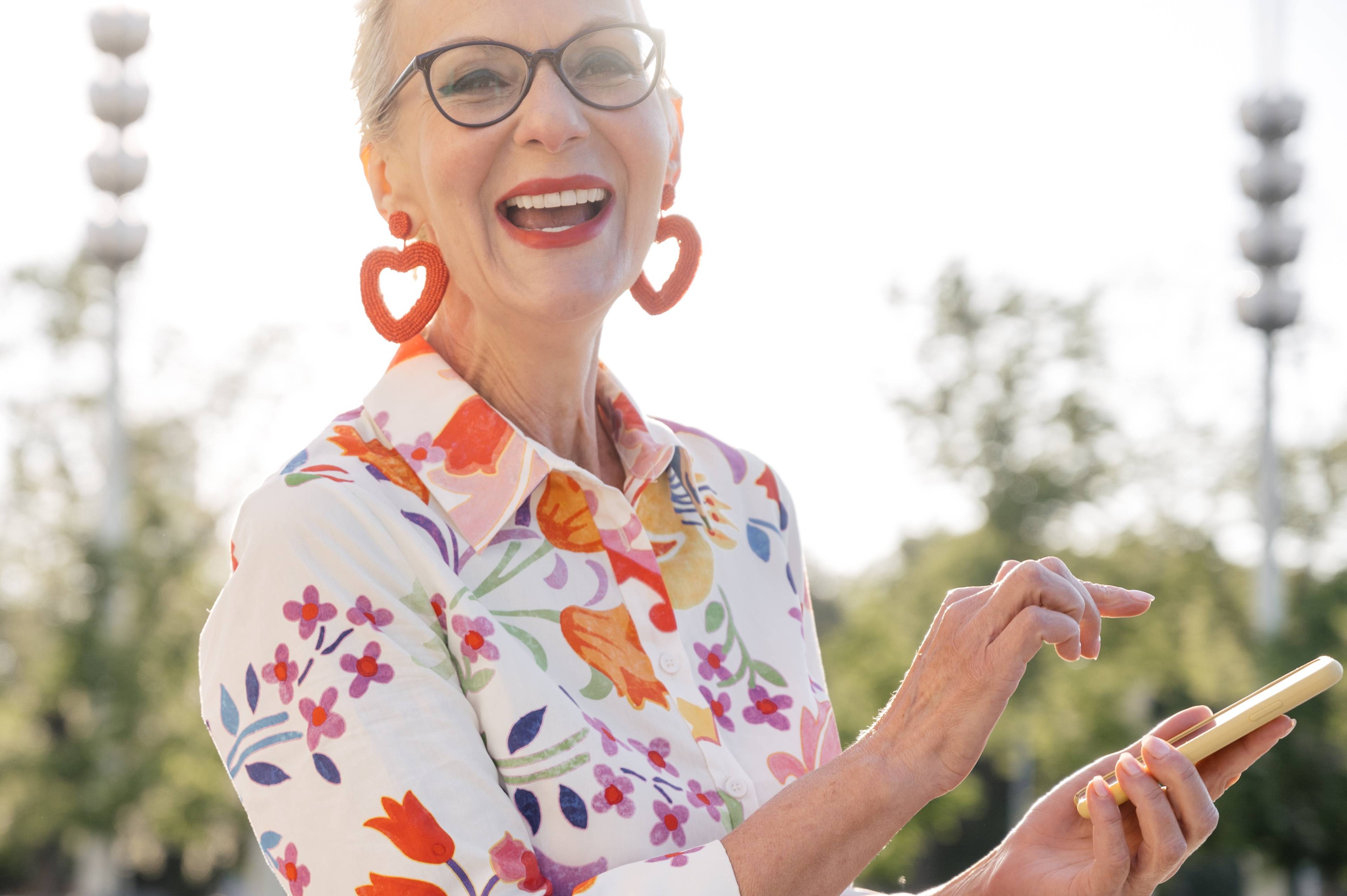 female wearing bright lipstick, earrings and blouse, thick framed glasses using phone and smiling outside in daylight garden
