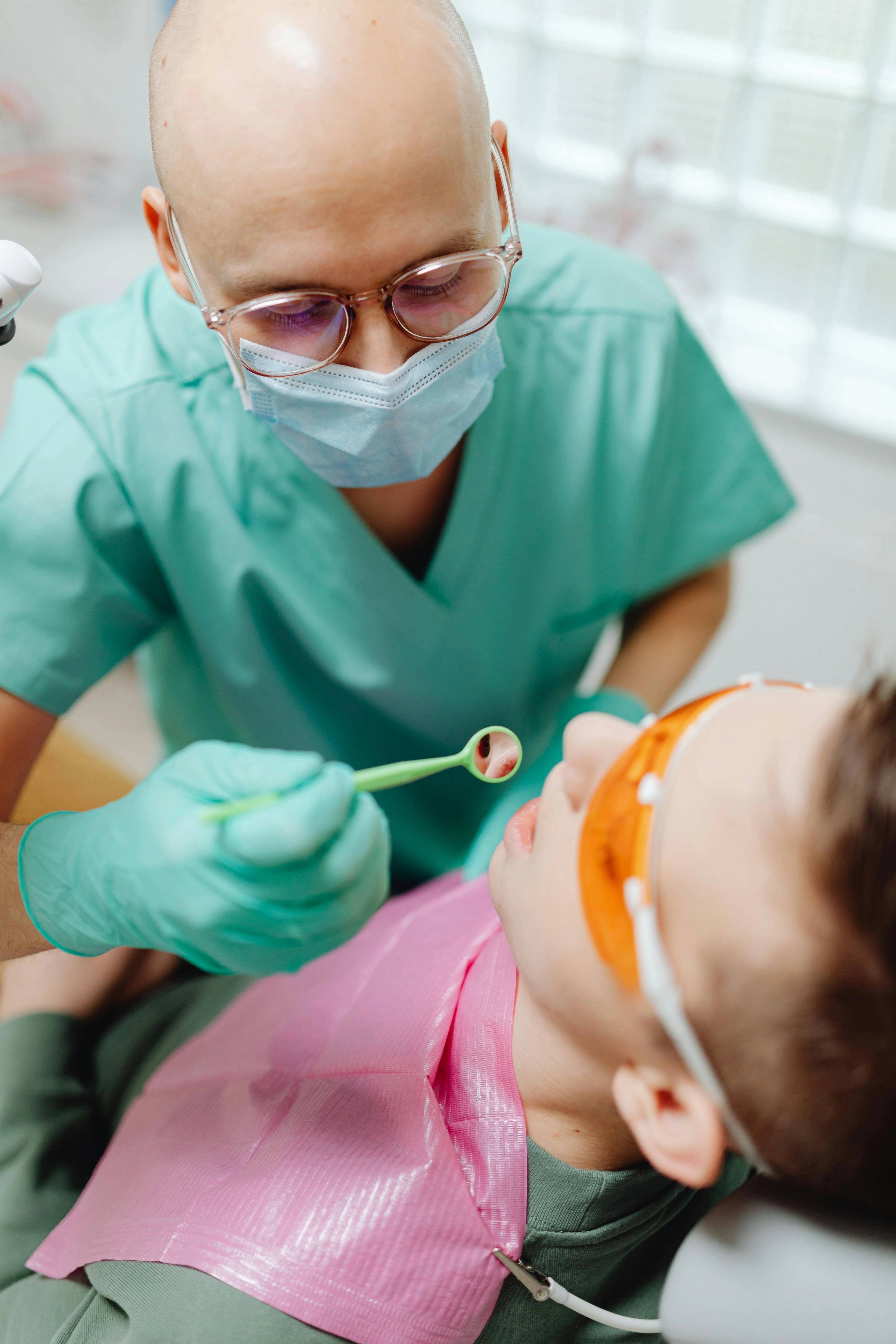male dentist in green uniform and mask holding small mirror in child's mouth while child wears eye protection
