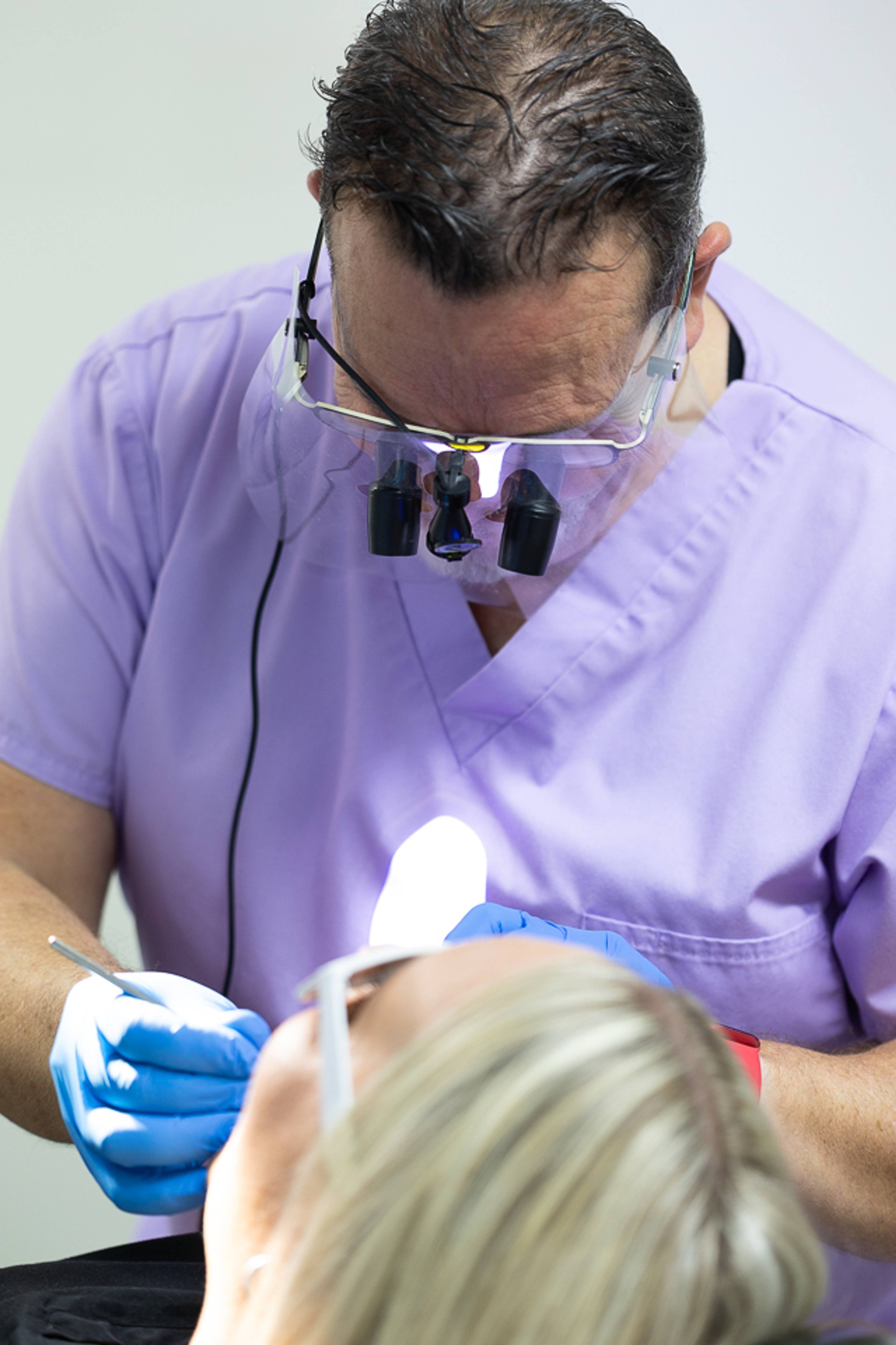 dentist in purple scrub is wearing loops and glasses whilst treatment patient in the chair in a clean calm surgery
