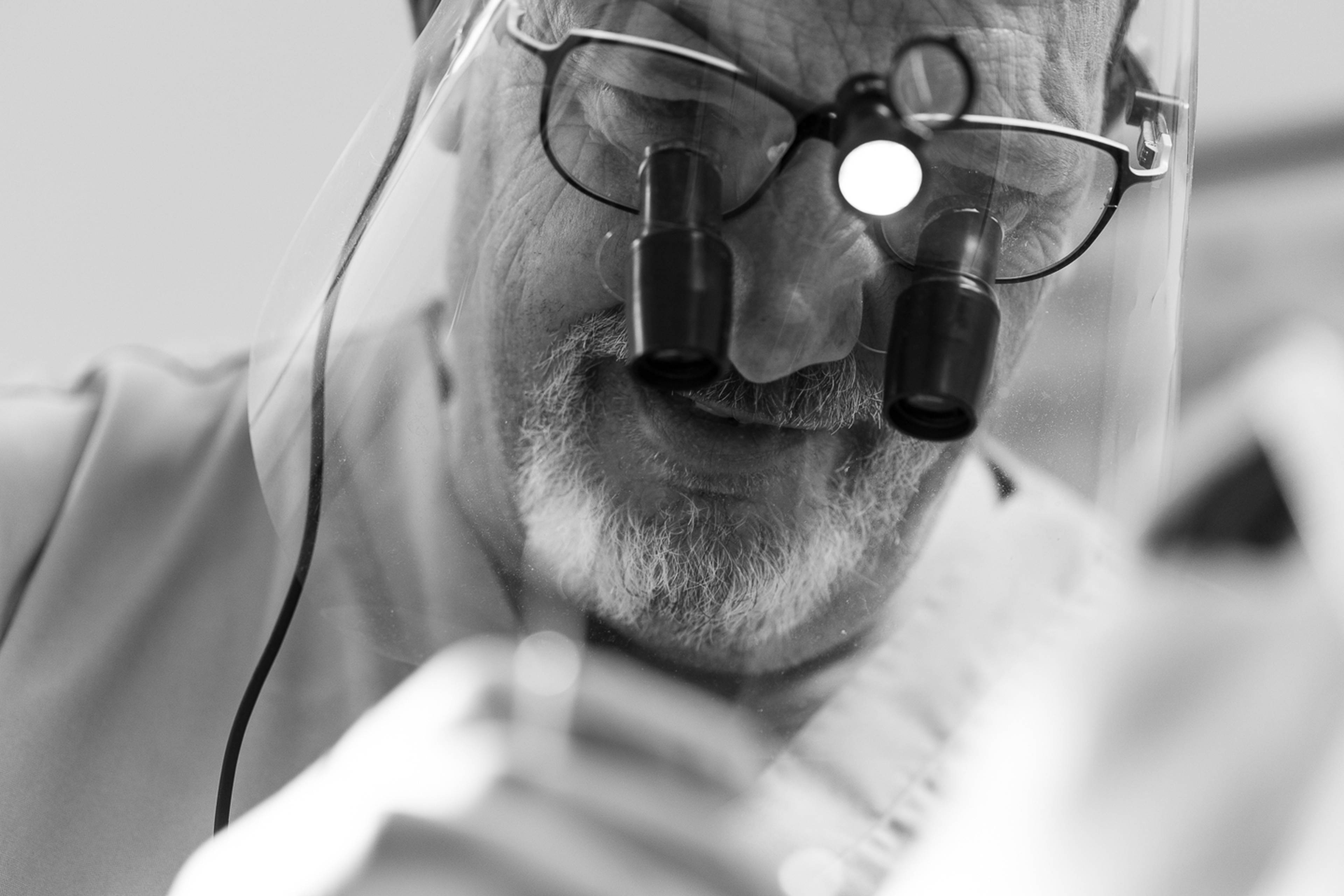 Black and white photo of a dentist wearing visor and loops looking down to treat a patient 