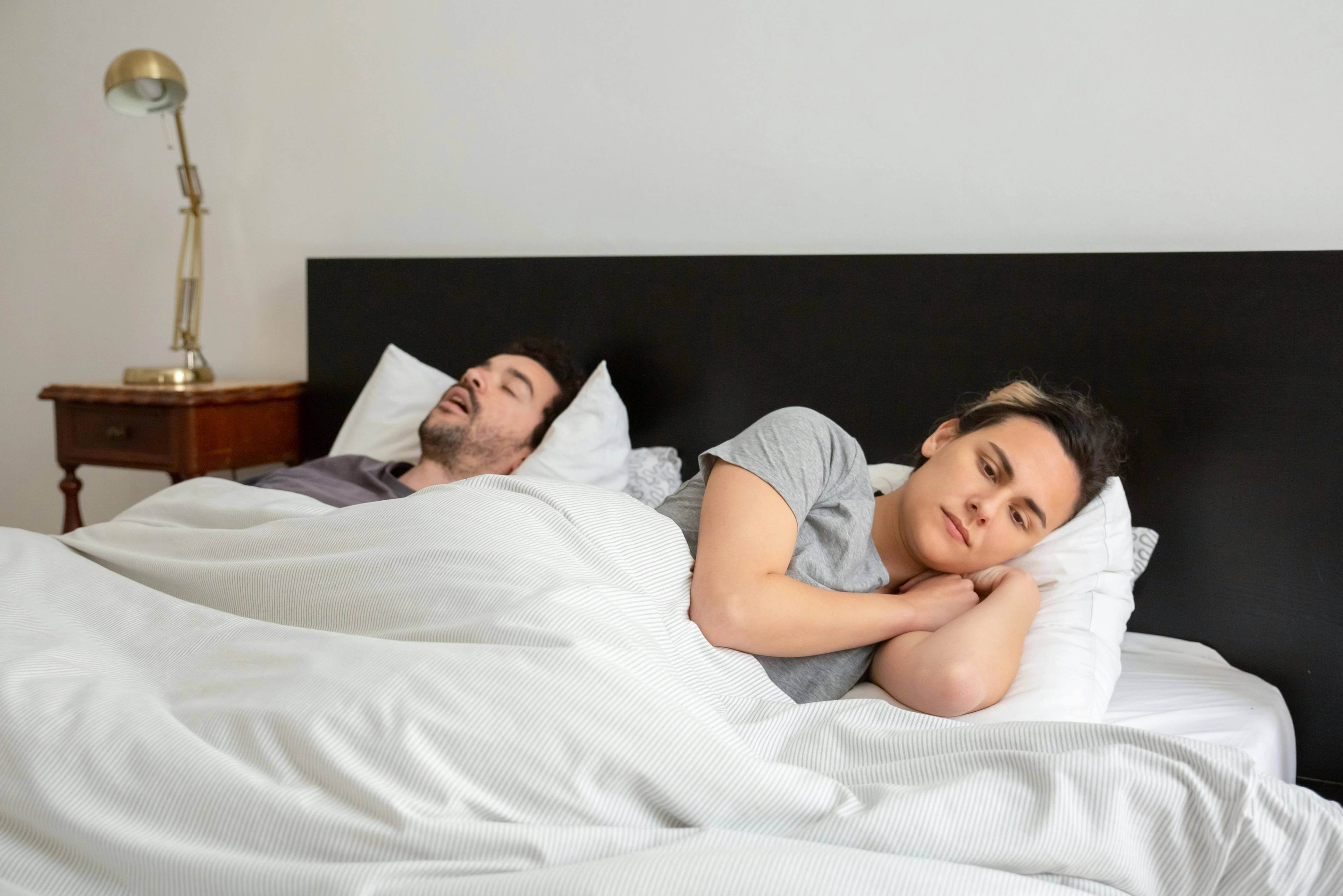 man snoring in bed with white sheets beside woman who is awake