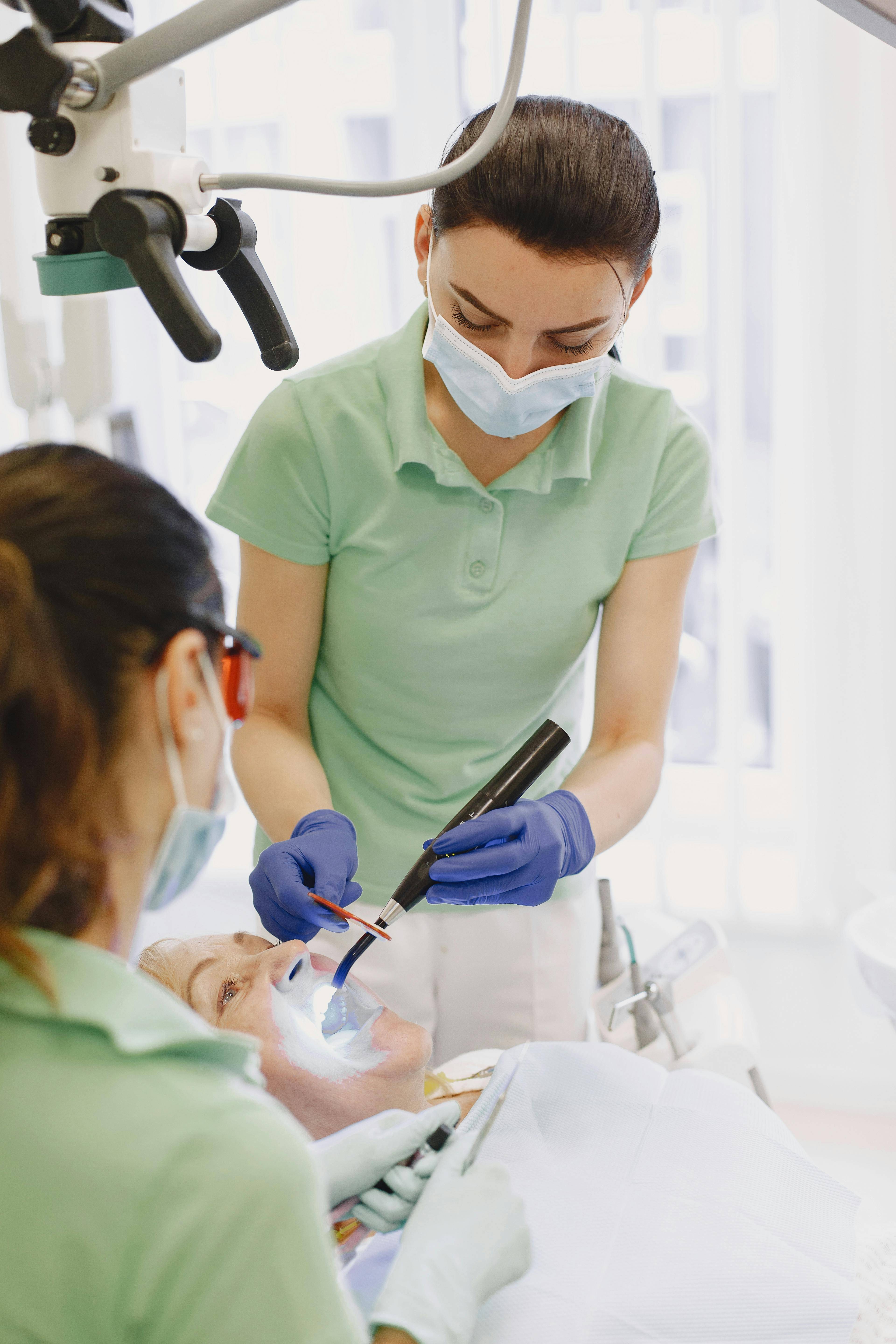 two female dentists wearing green uniforms, blue gloves and face masks treating a patient in surgery