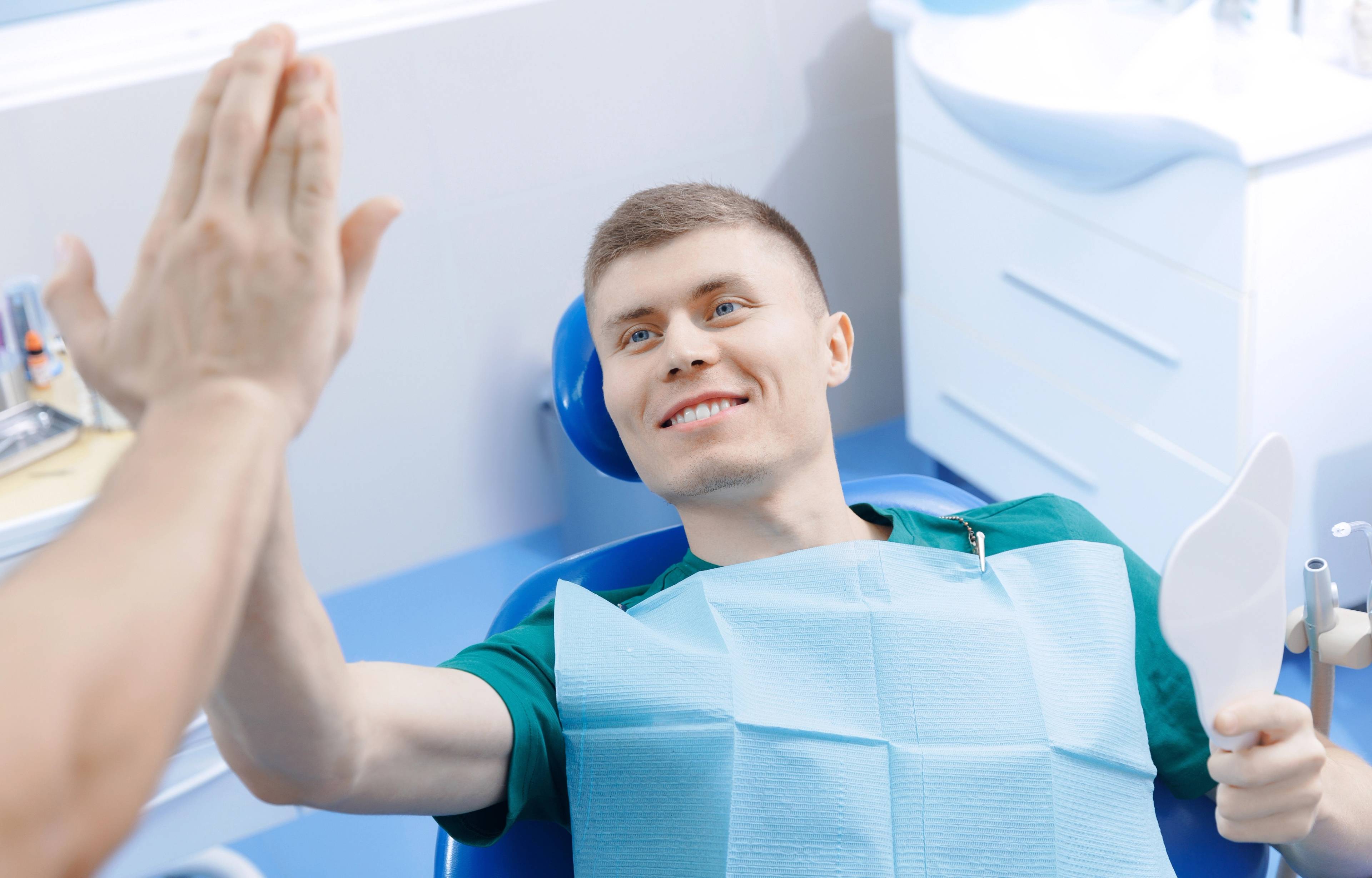 male patient smiling in dental chair holding handheld mirror and high-fiving dentist 