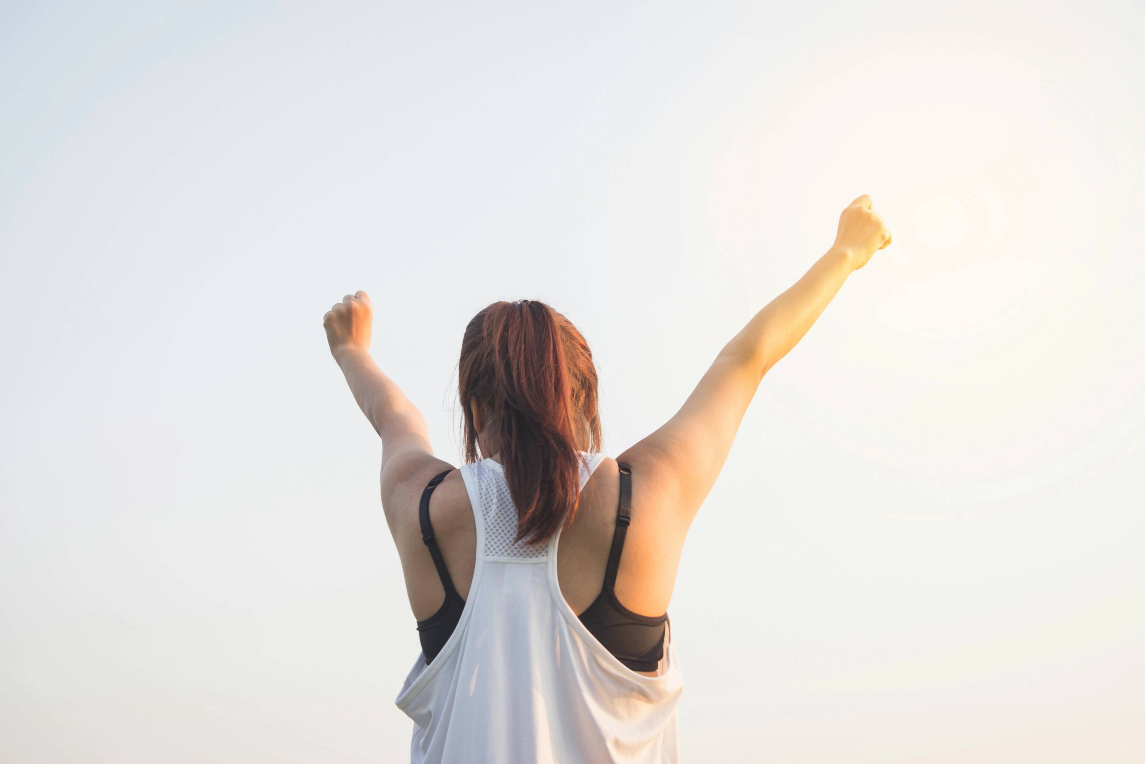 female standing outside under the sun with both arms raised 