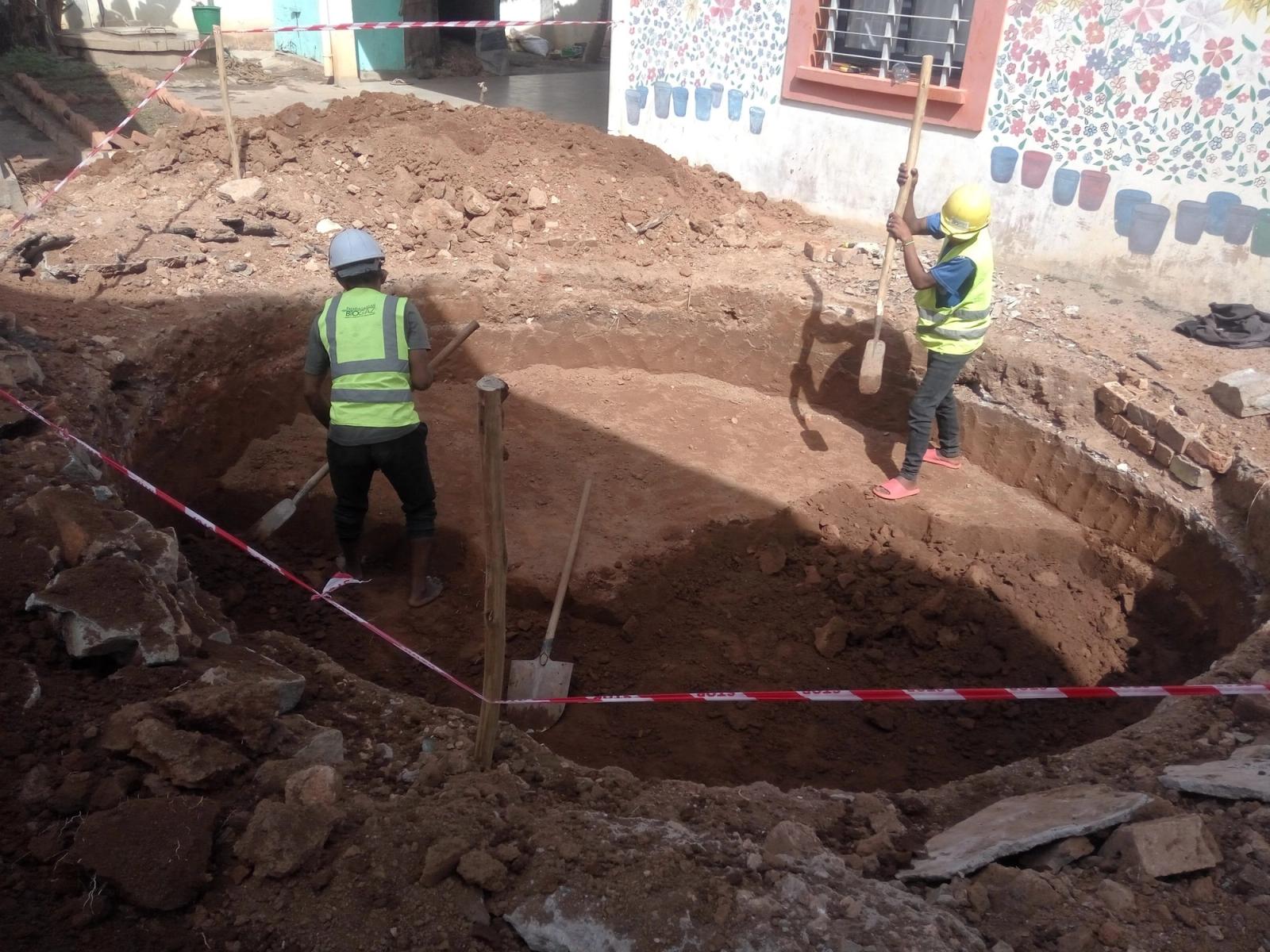 Zwei Personen in Warnwesten arbeiten mit Schaufeln an einer Baugrube.