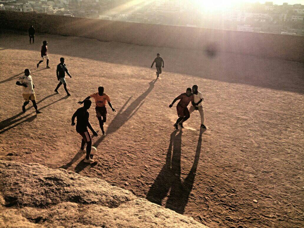 Kinder spielen Fußball auf einem Sandplatz im Abendlicht.