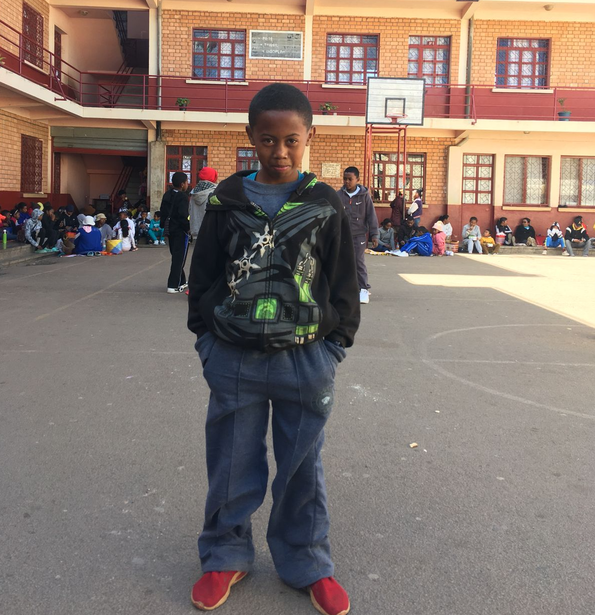 Ein Kind steht im Schulhof vor dem Gebäude; weitere Kinder sitzen im Hintergrund.