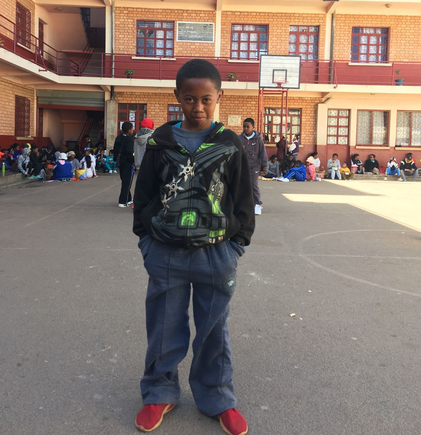 Ein Kind steht im Schulhof vor dem Gebäude; weitere Kinder sitzen im Hintergrund.