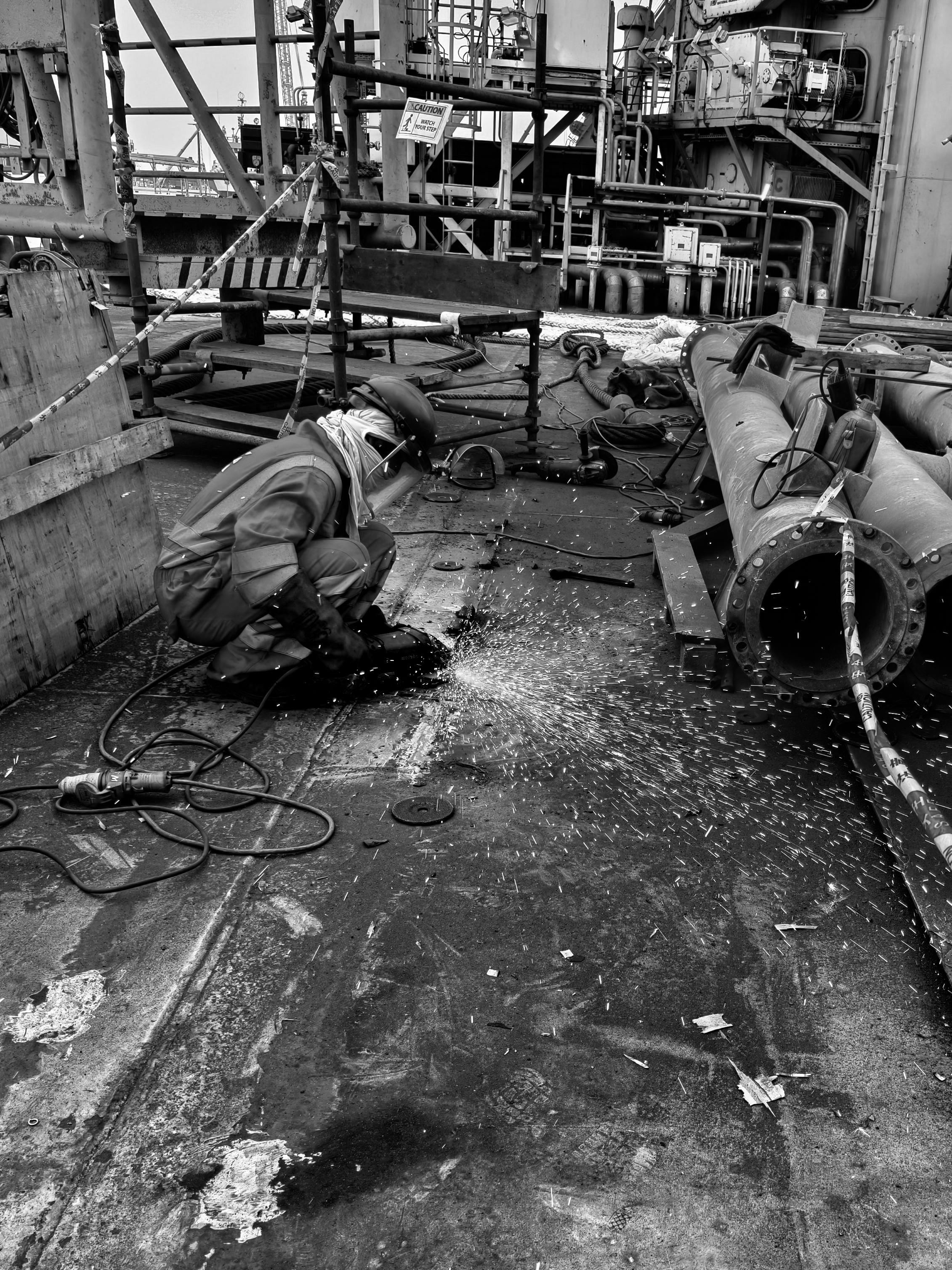 Worker grinds metal on an industrial deck, sending sparks across pipes and equipment.