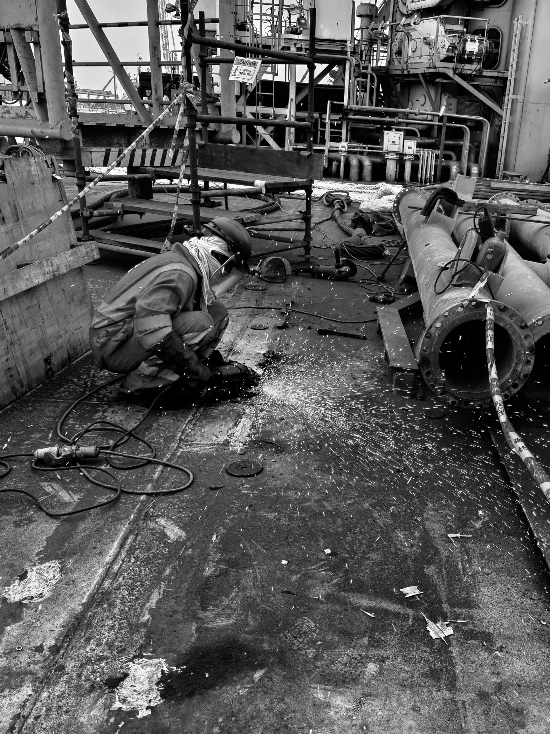 Worker grinds metal on an industrial deck, sending sparks across pipes and equipment.