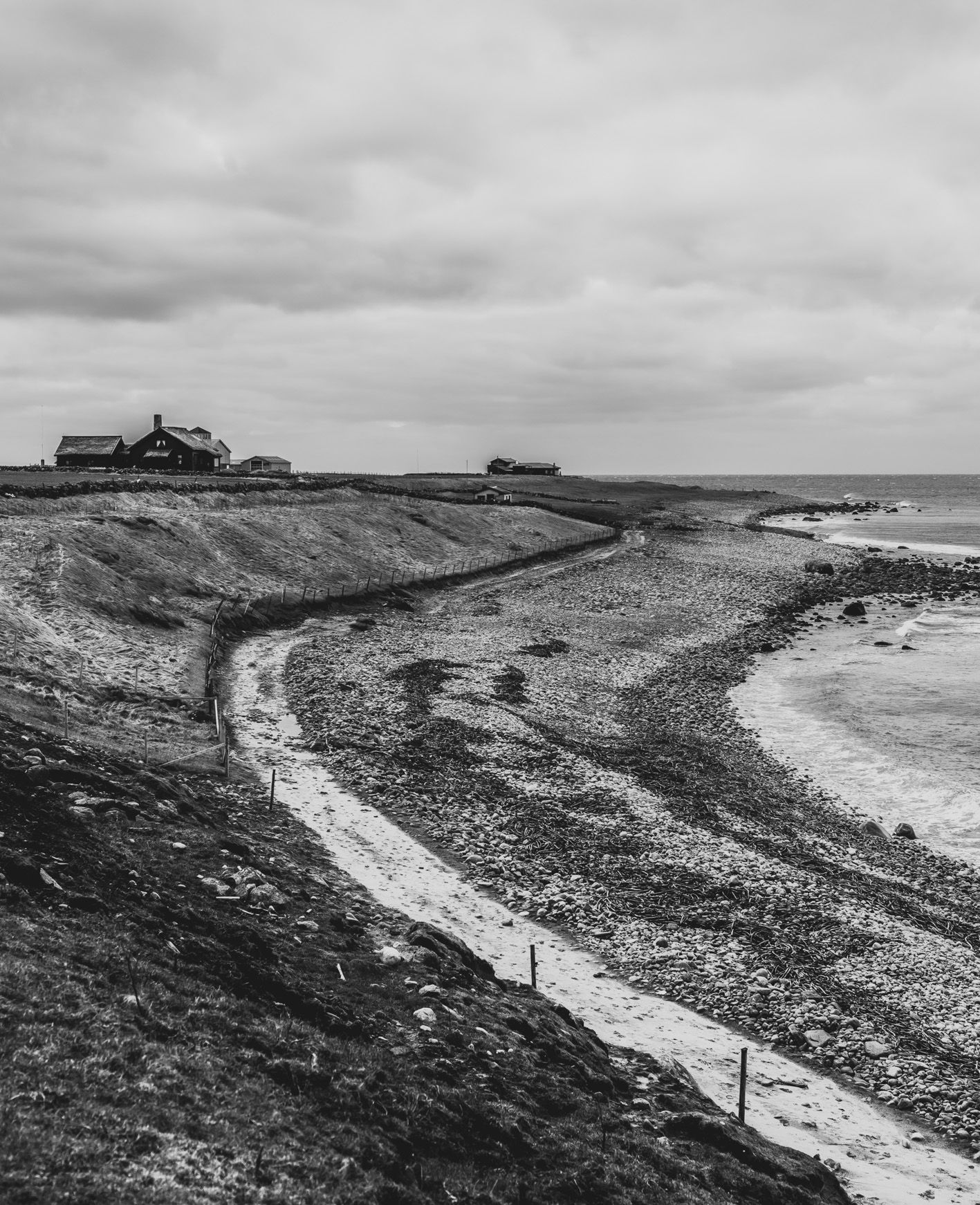 Black-and-white view of a rocky coastline with a dirt path and distant seaside houses