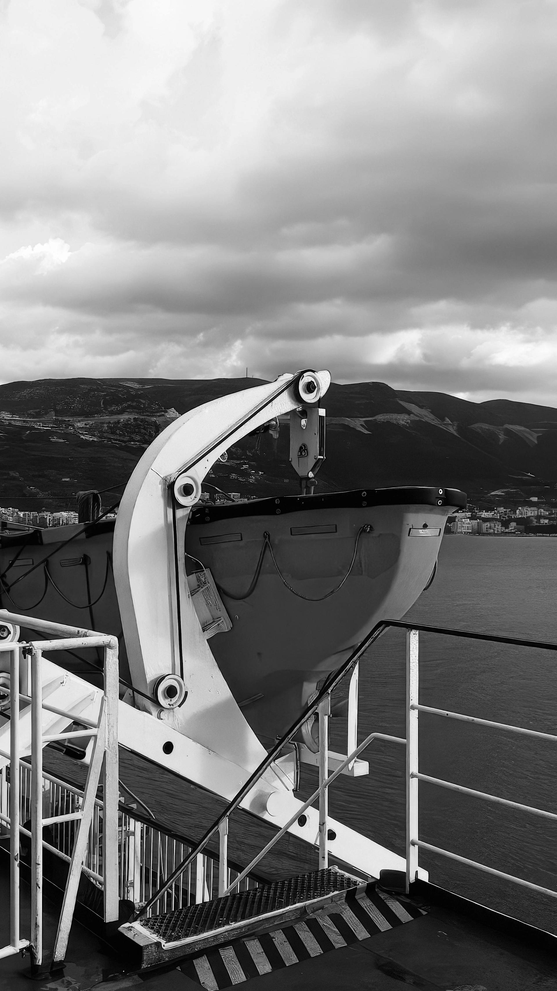 Black-and-white view of a ship’s lifeboat on deck with mountains and sea behind
