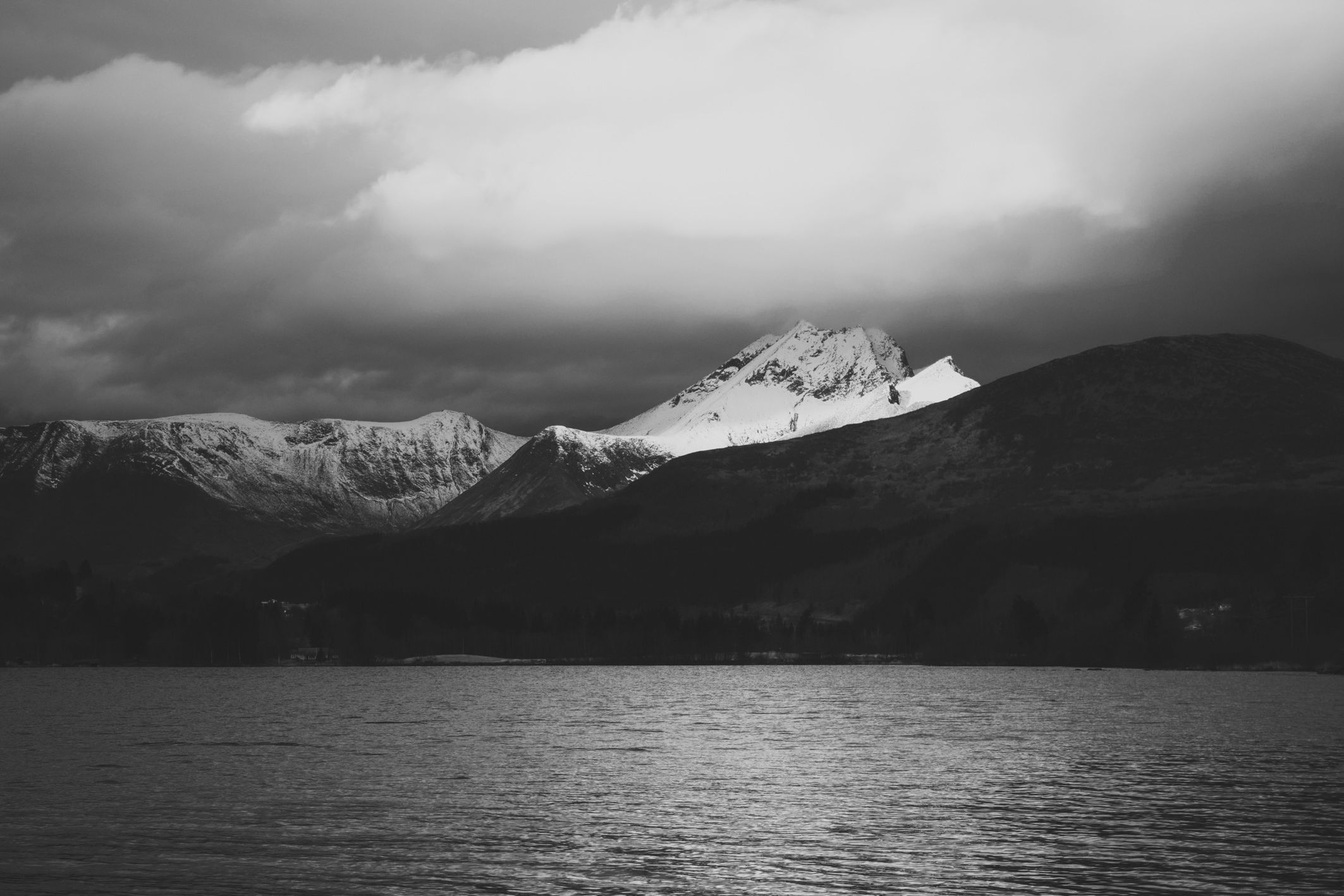 Snow-capped mountain range under heavy clouds above a dark lake in black and white