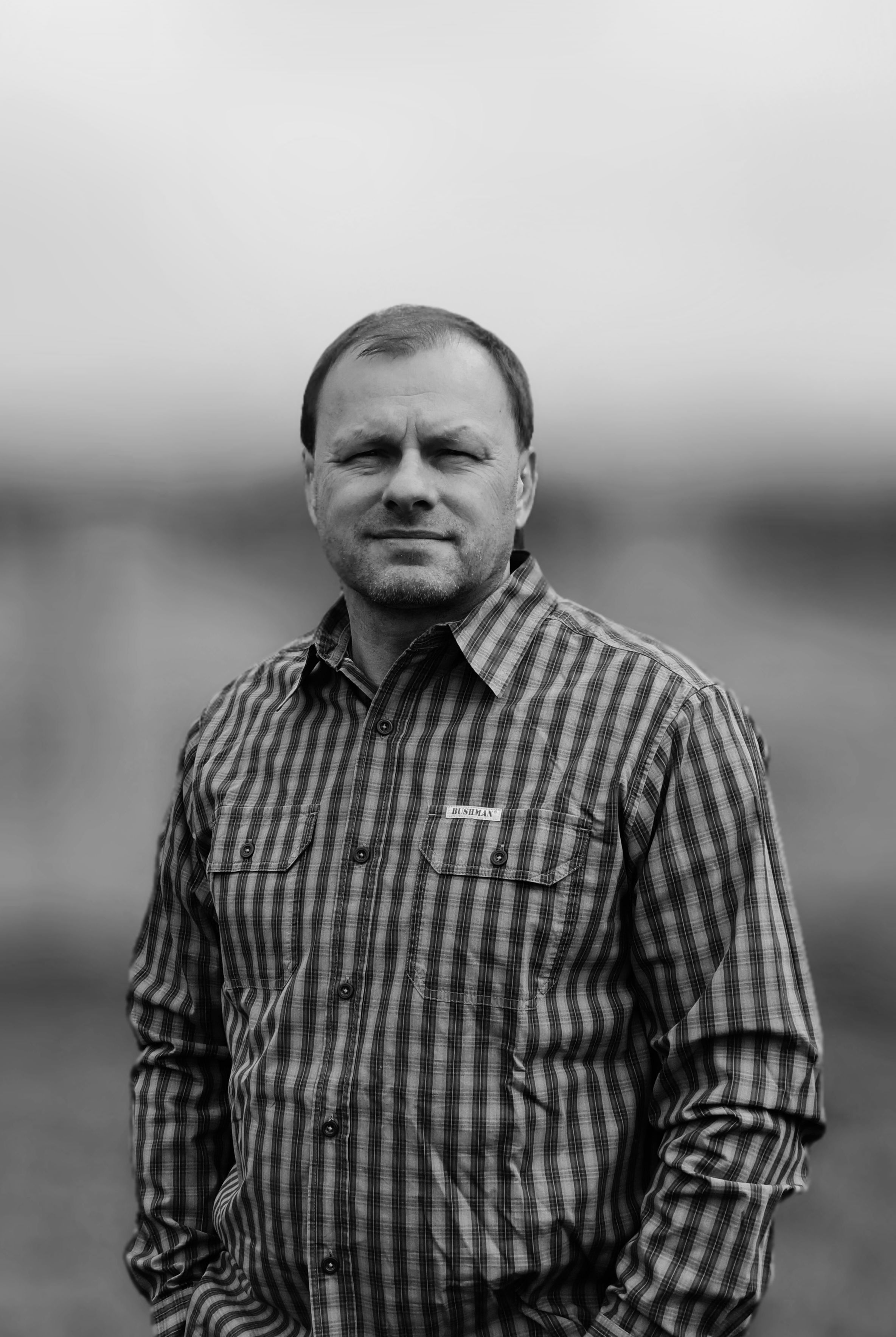 Man in a plaid shirt standing outdoors, looking at the camera in black and white