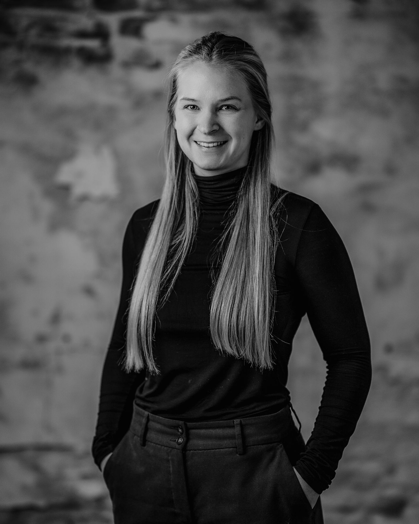 Smiling person with long straight hair in black clothes, hands in pockets, studio portrait