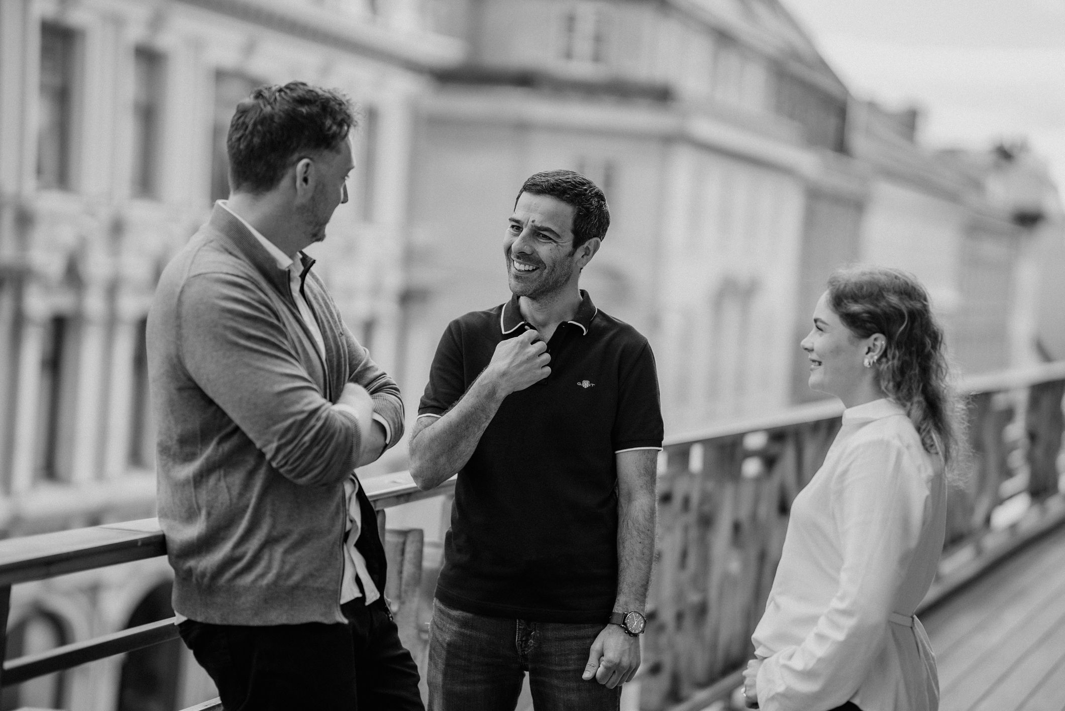 Three people stand on a balcony, smiling and talking together in a city setting.