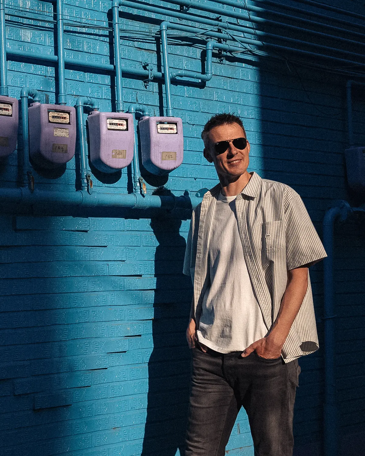 Photo of Jitse walking into a patch of light in front of a blue wall, boasting sunglasses