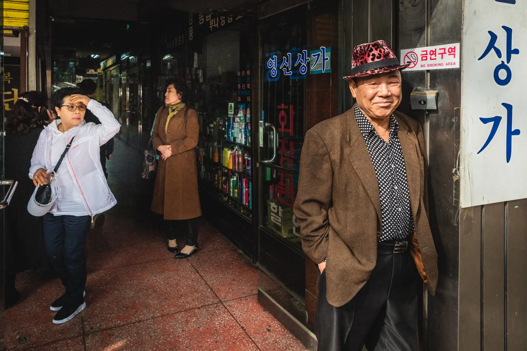 A Korean man in a pink leopard-print hat stands at the entrance to an alley as two women stand behind him