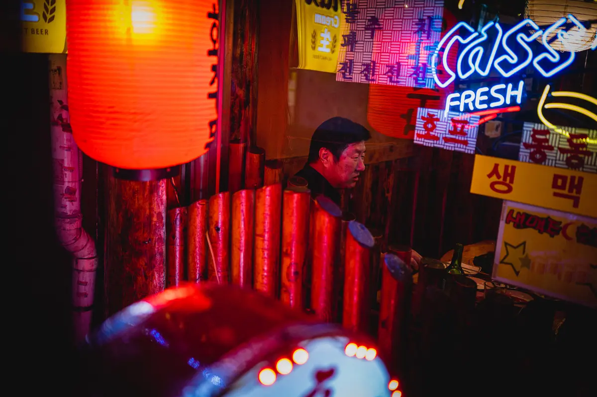 A middle-aged Korean man sits at a neon-lit bar with Korean text and beer logos in Jongno, Seoul
