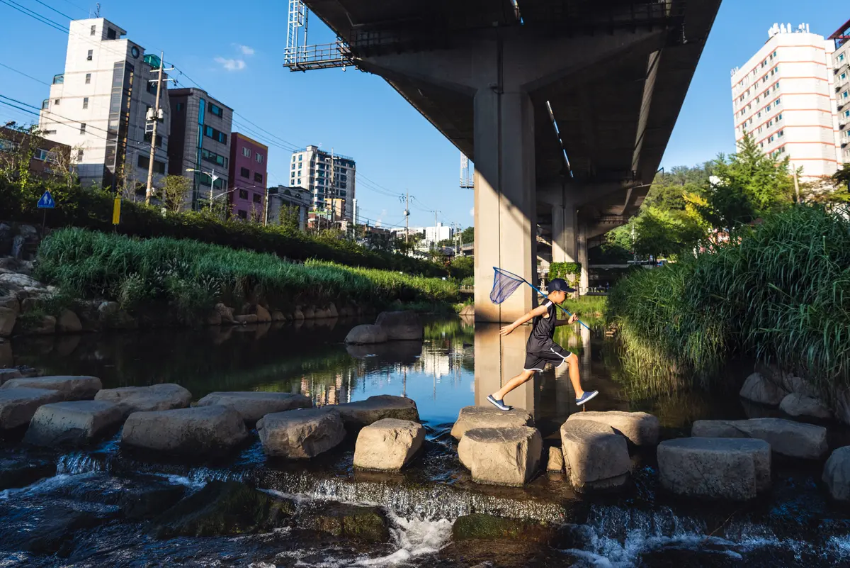 Young Korean boy runs across a river path under a bridge holding a fishing net at the Hongjecheon stream in Seoul