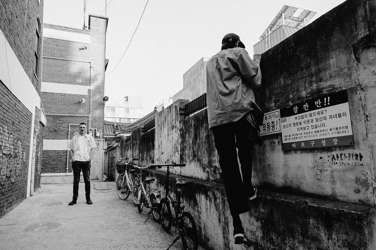 Black and white photo of Jitse standing still and Chris trying to climb a stone wall