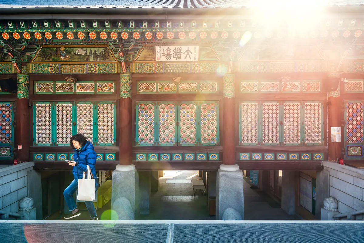 A woman looks at her smartphone while she walks up the steps at a Buddhist temple as sunlight flares from above in Seoul