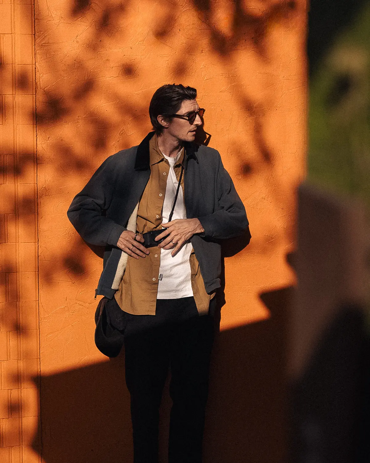Portrait of Chris standing in afternoon sunlight, in front of an orange wall with shadow patches