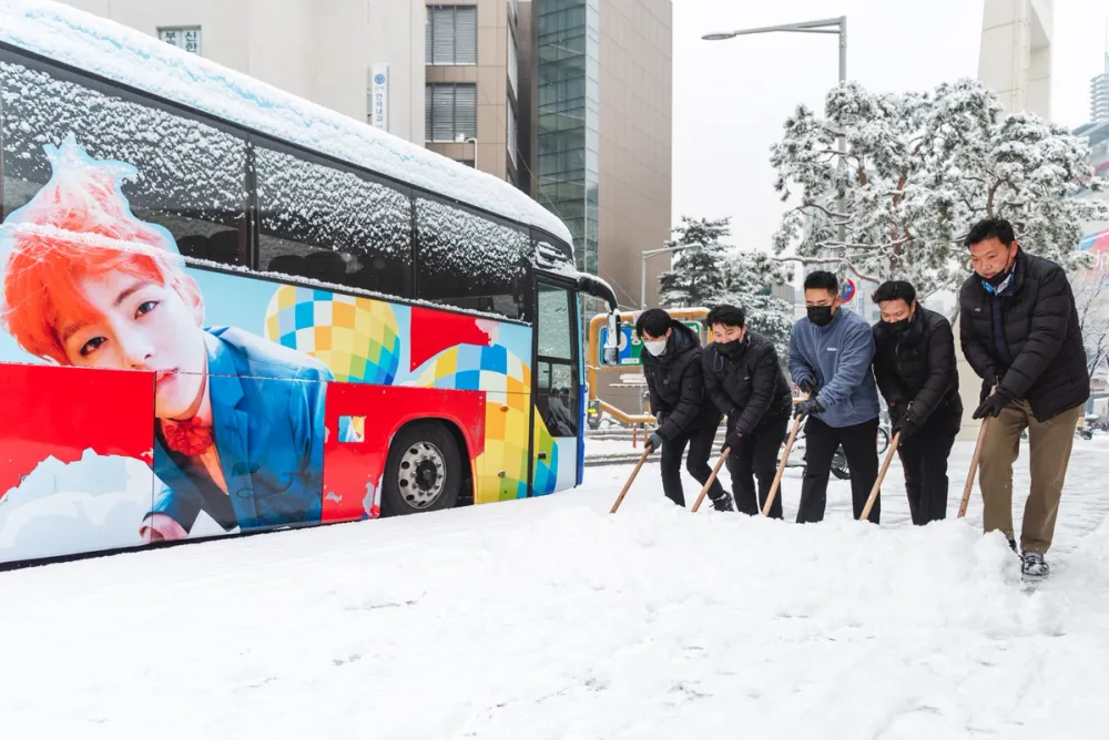 Men clearing snow from a Seoul street beside a colourful bus wrapped in a K-pop advertisement, winter.