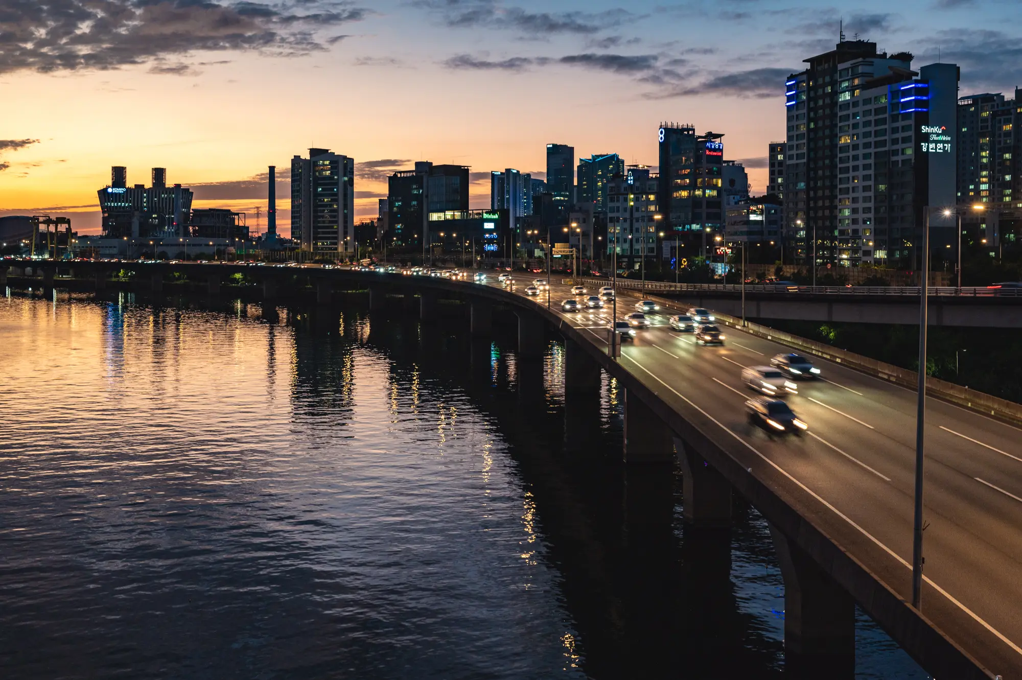 Traffic flows at dusk alongside the Han River as tall buildings are silhouetted in the background