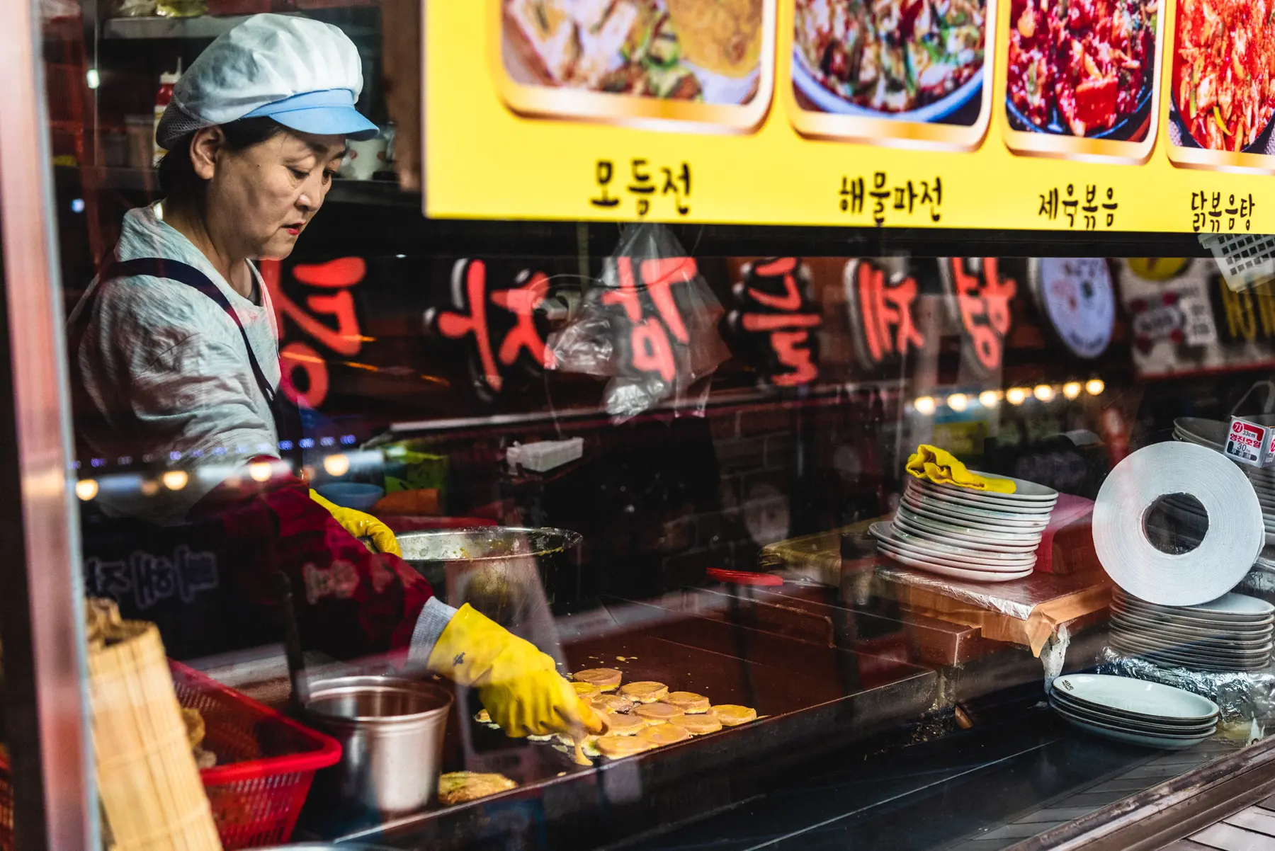 A Korean woman wearing a chef's uniform makes jeon behind a restaurant's glass window
