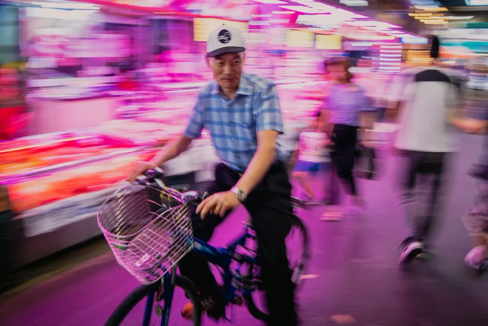 A man cycling through a busy Seoul market, the motion blur of neon-lit stalls and pedestrians surrounding him.