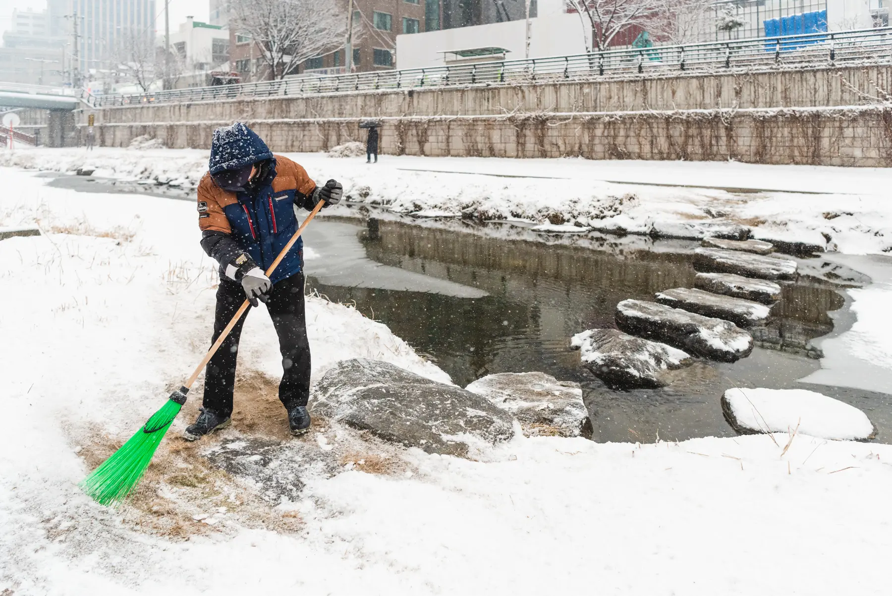 A Korean man sweeps snow with a green broom by a river while snow falls in Seoul, South Korea
