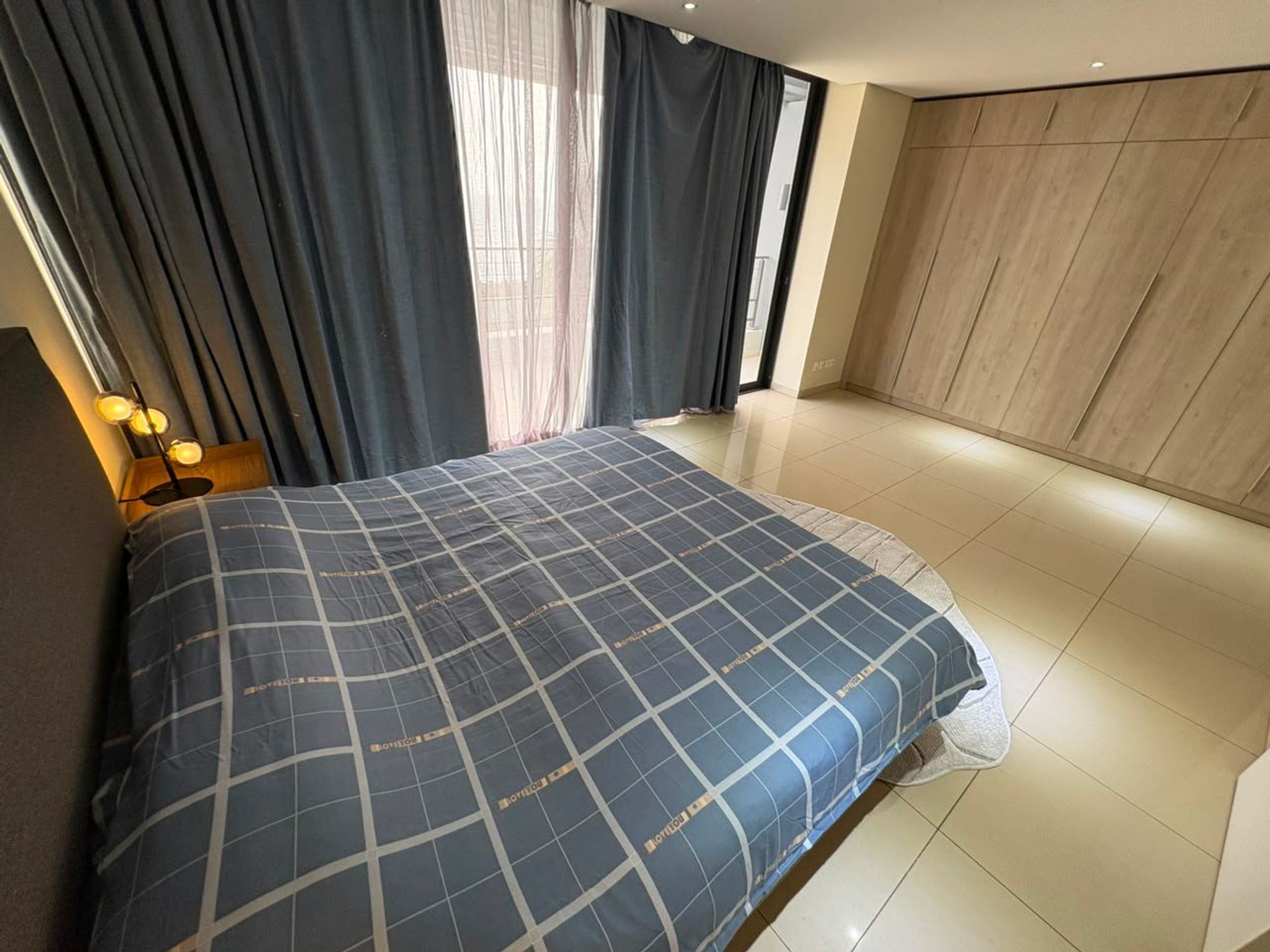 Chambre du duplex meublé à Mermoz avec lit à couette à carreaux, rideaux gris et grande armoire en bois