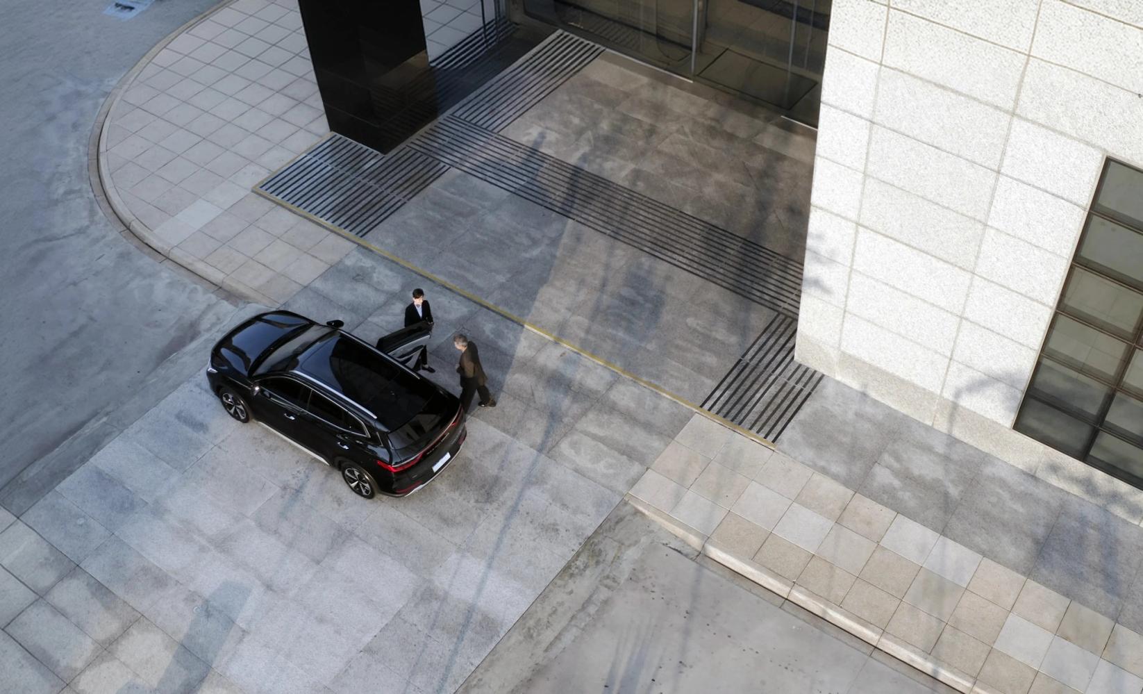 Um SUV blindado preto visto de cima, com o chofer segurando a porta traseira aberta para um passageiro