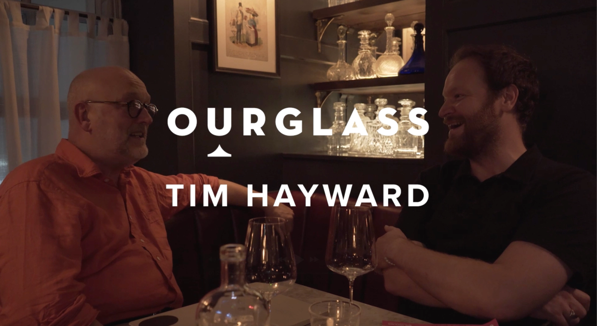 Tim Hayward and Benedict Johnson chat over wine in a dimly lit corner of Noble Rot, surrounded by glassware, during the filming of the Ourglass Tapes video series.
