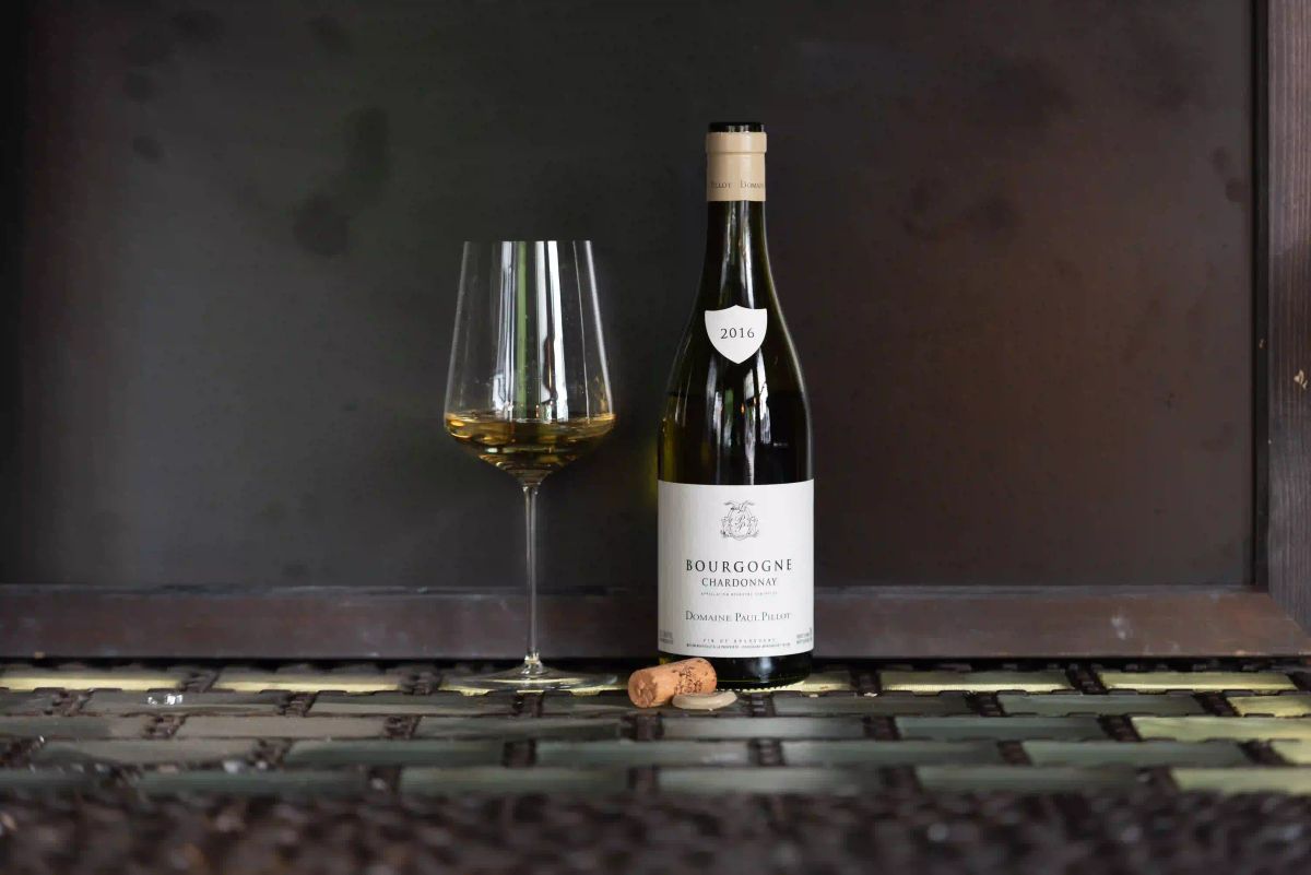 A half-full glass of white Burgundy beside an opened 2016 Bourgogne Chardonnay from Domaine Paul Pillot, two corks resting on a textured bamboo surface, low-light editorial still life.