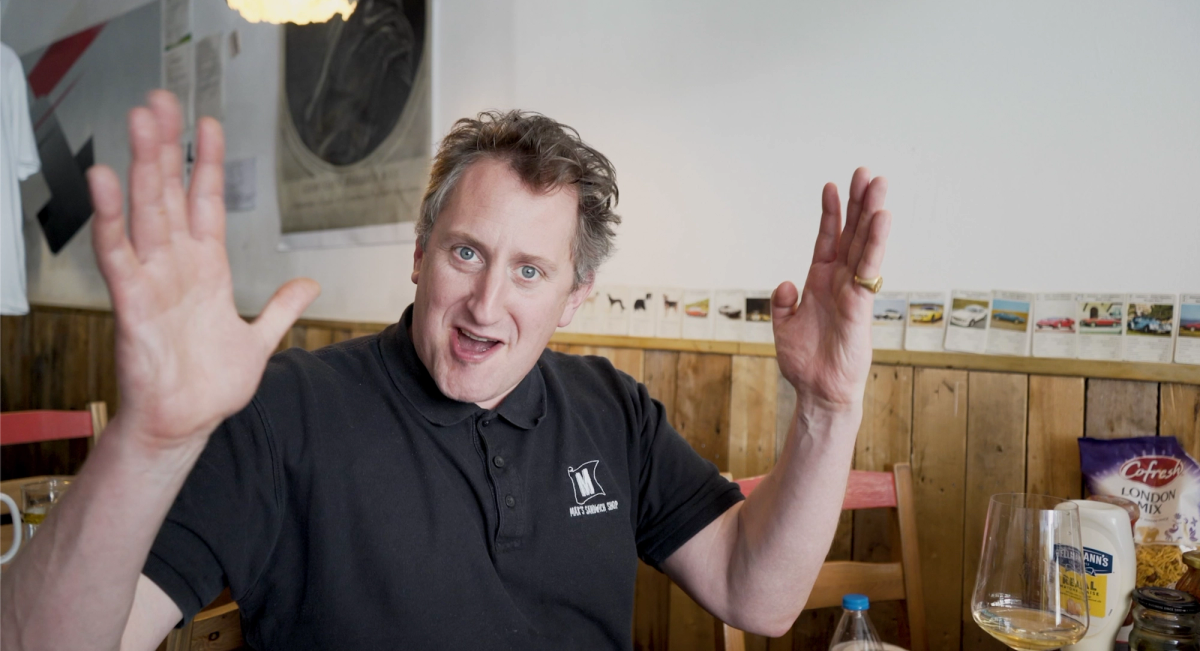 Max Halley gesturing enthusiastically at Max’s Sandwich Shop in London, with a wine glass and condiment jars in the foreground, shot during the Ourglass Tapes video series on wine and food pairing.