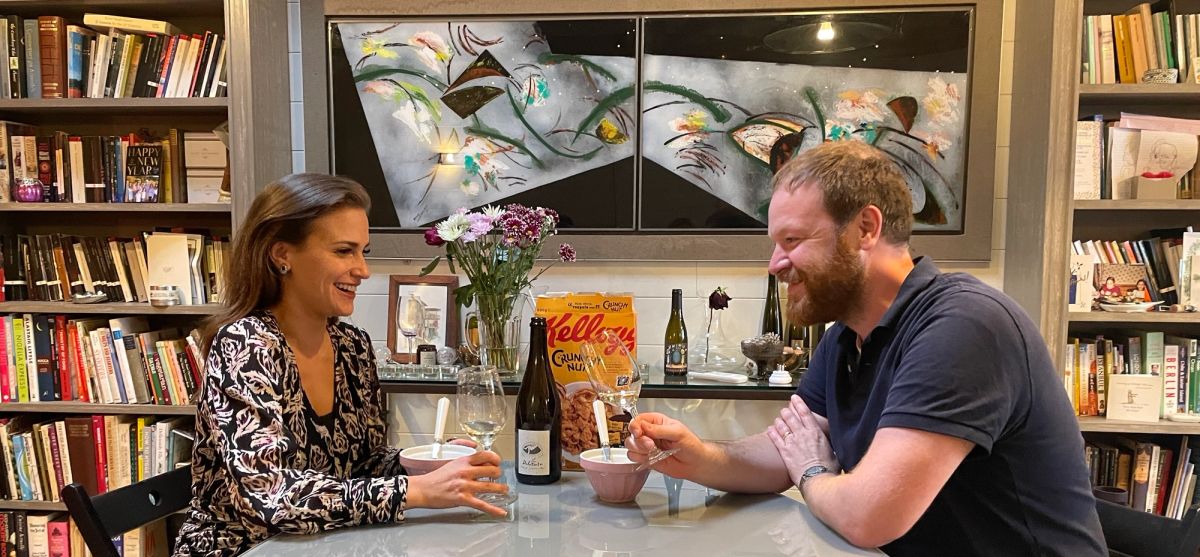 Amelia Singer and Benedict Johnson sit at a table smiling over bowls of Crunchy Nut Cornflakes with wine glasses, a bottle of La Dilettante by Catherine and Pierre Breton, and a box of cereal between them, set against bookshelves and modern art.