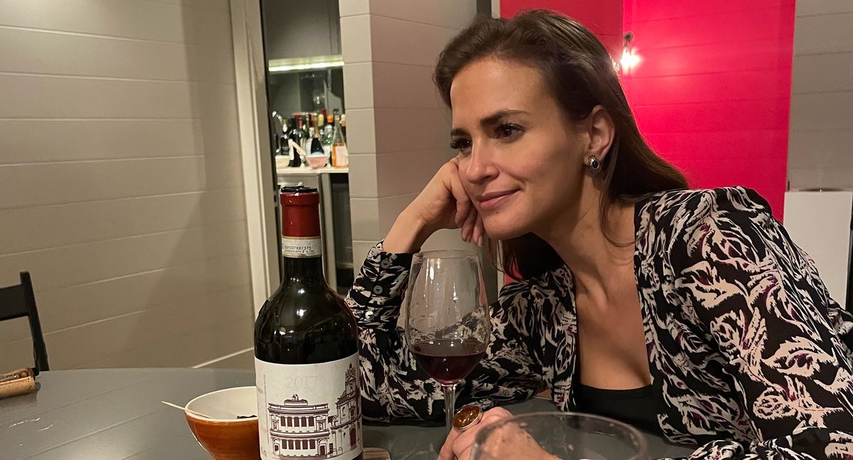Amelia Singer rests her chin on her hand while enjoying a glass of COS Cerasuolo di Vittoria, seated at a table with the bottle, slices of cured meat, and two other wine glasses, with a vivid red backdrop behind her.