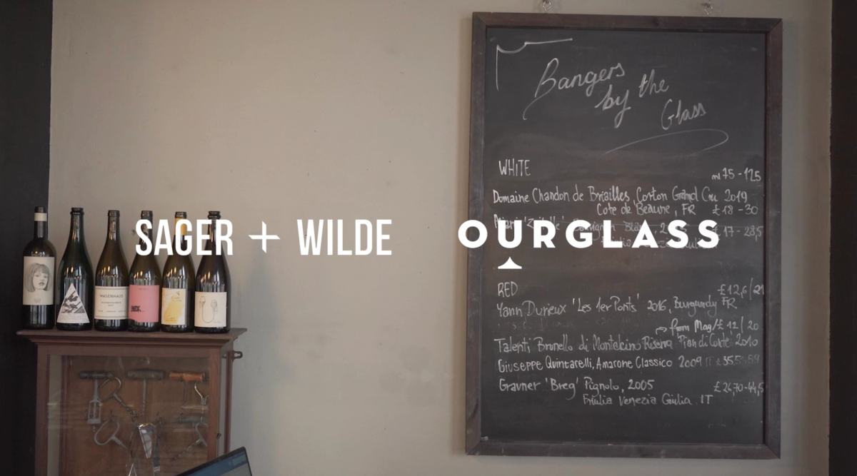 A blackboard wine list titled “Bangers by the Glass” at Sager + Wilde, with a row of wine bottles displayed beside it and the Sager + Wilde and Ourglass logos overlaid.