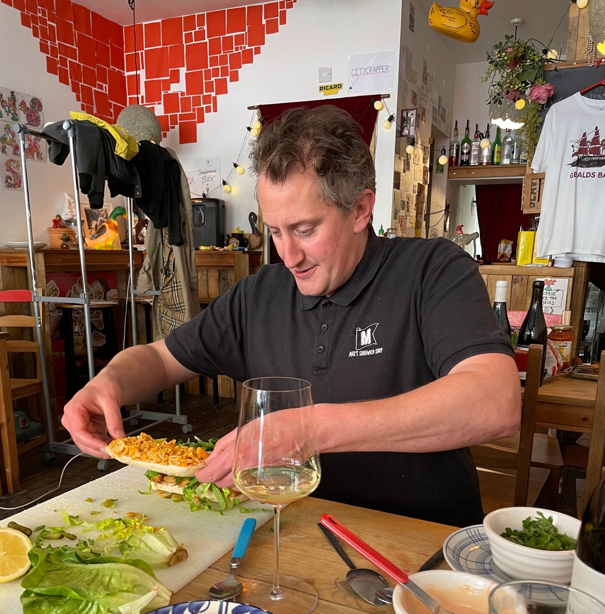 Max Halley assembles a sandwich at Max’s Sandwich Shop, surrounded by vibrant decor, a glass of white wine, and ingredients laid out on a wooden table.
