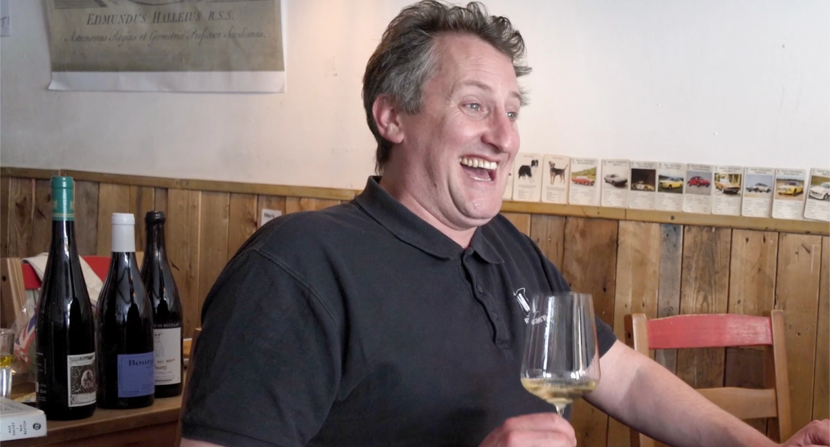 Max Halley laughing mid-conversation while holding a glass of white wine at Max’s Sandwich Shop, with bottles lined up behind him and vintage car cards pinned along the wall.