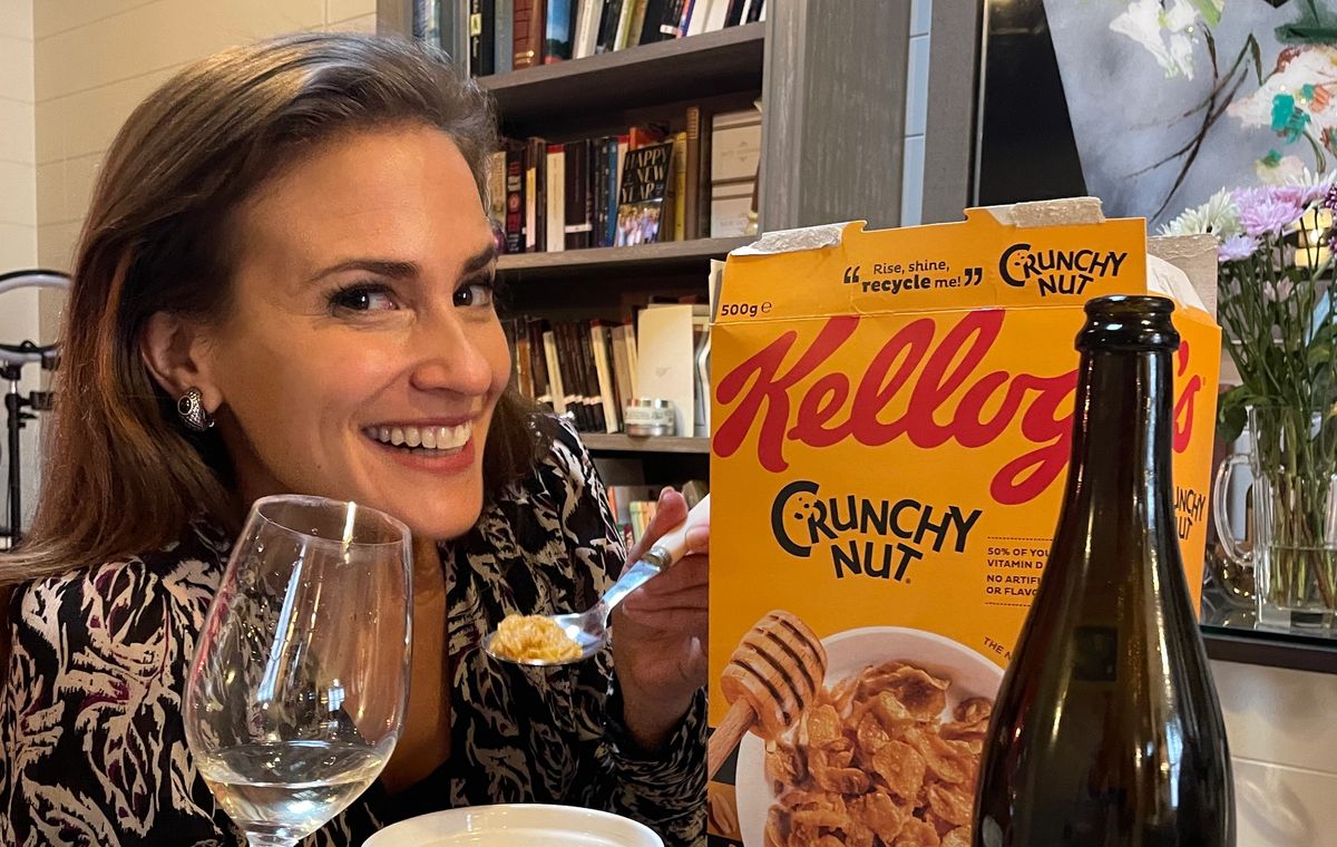 Amelia Singer smiles while holding a glass of wine and a spoonful of Crunchy Nut Cornflakes, seated beside a cereal box and a bottle of La Dilettante wine.