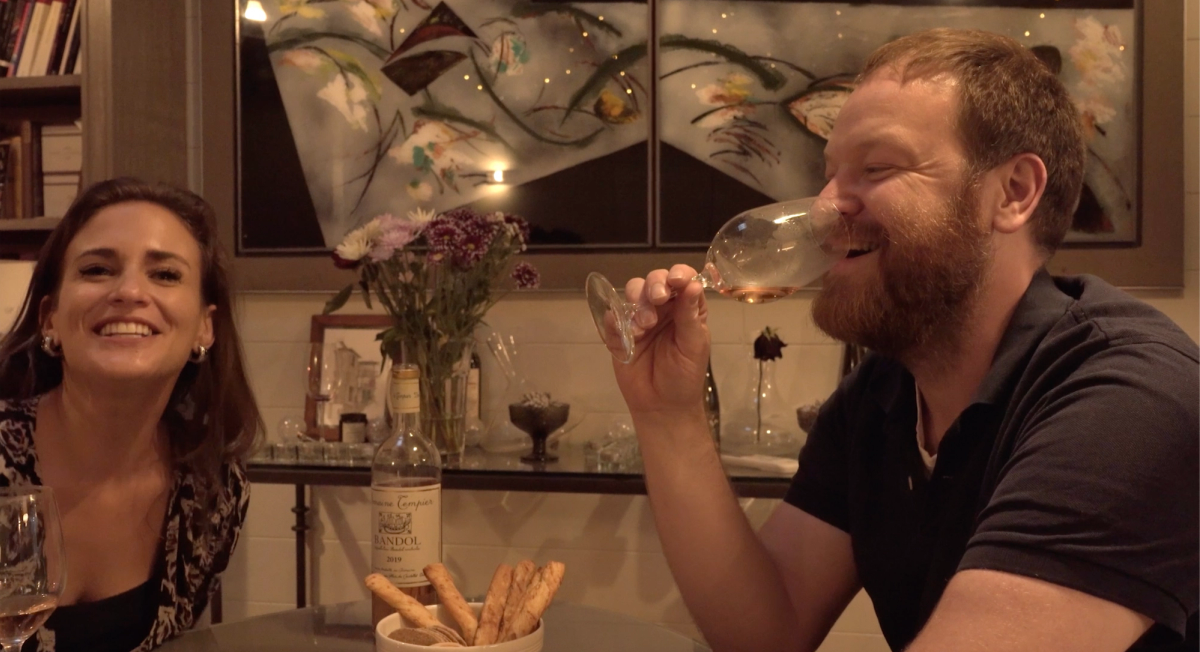 Amelia Singer and Ben share a laugh at the table while tasting Bandol Rosé from Domaine Tempier, with a bottle of wine and breadsticks in front of them and abstract art and flowers in the background.