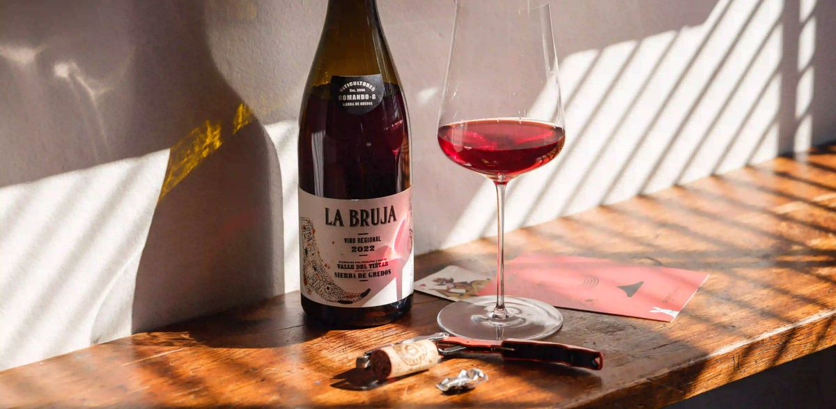 A glass of red wine beside an open bottle on a wooden table in natural light, ready to taste.