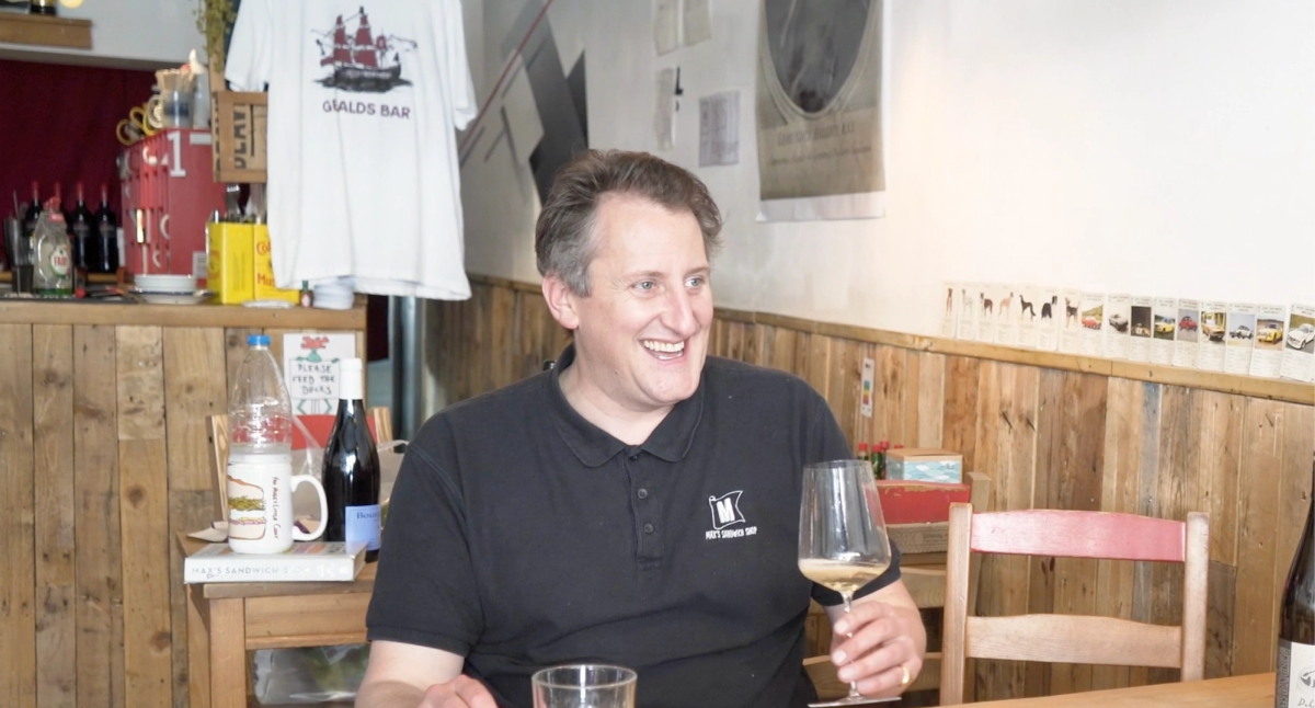 Max Halley at Max’s Sandwich Shop during filming of the Ourglass Tapes series, smiling mid-conversation with a glass of white wine. Wine bottles, ingredients, and his cookbook frame the scene.