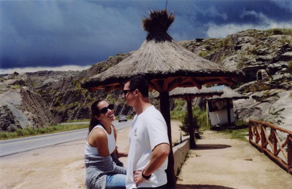 Two people talking under a thatched shelter beside a mountain road