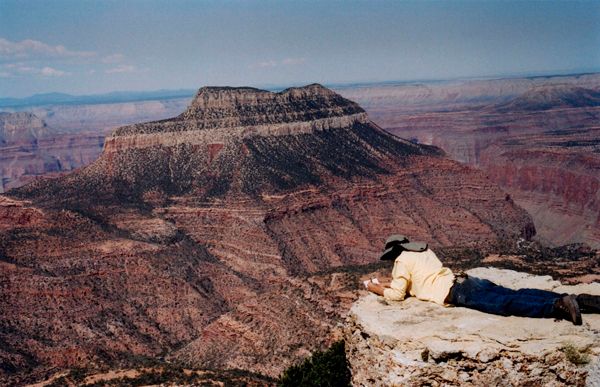 Person lying on cliff edge sketching a distant flat-topped mesa